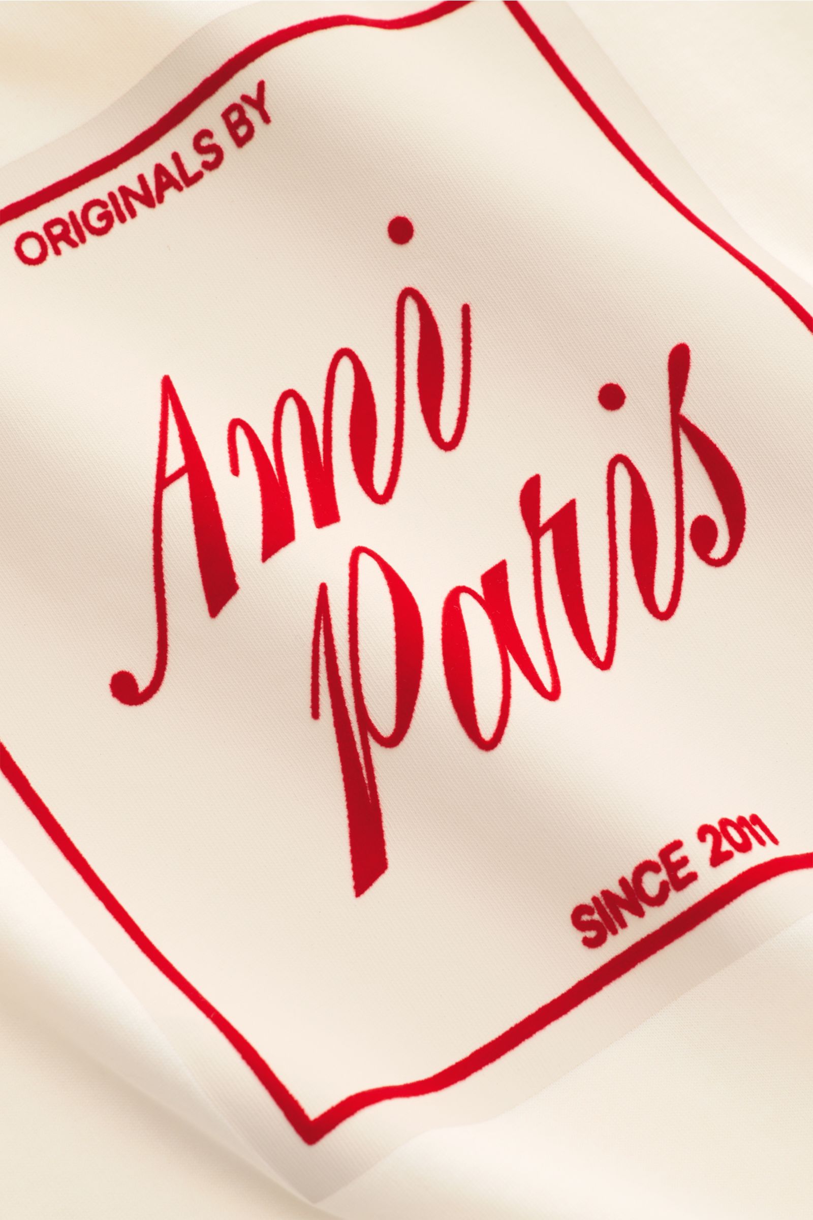 Nahaufnahme des Ami Paris Rundhals-T-Shirt creme, gefertigt aus reiner Baumwolle mit geflocktem roten AMI PARIS-Schriftzug frontal, weicher Griff, Regular Fit, Rundhalsausschnitt und rückwärtigem Logo-Stitching.