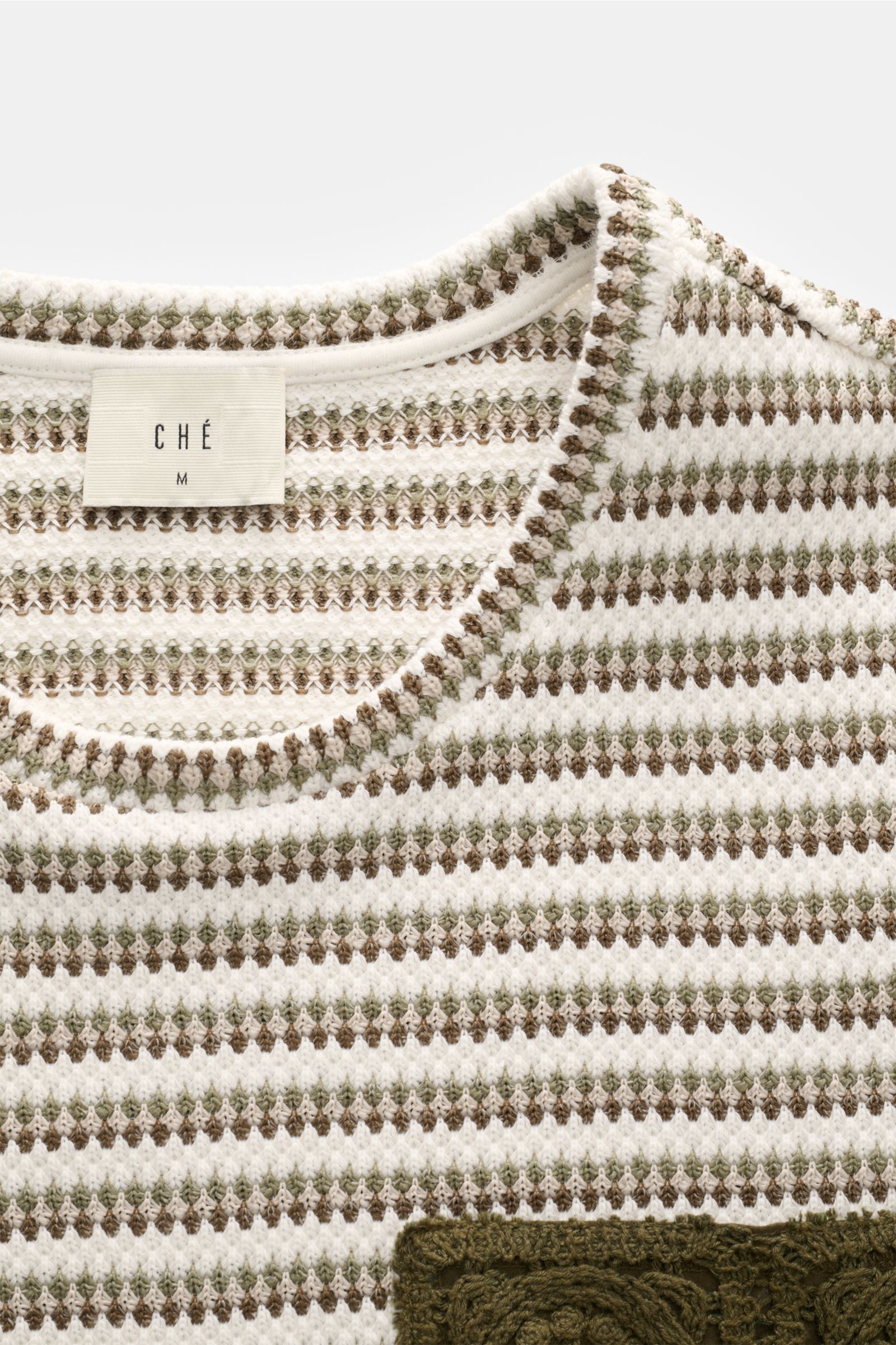 Close-up front view of Ché Kurzarmpullover 'Mathias' creme/salbei/dark olive gestreift with fine striped crochet knit, round neckline, and chest pocket.  

Lässiger Urlaubslook: Der Kurzarmpullover von CHÉ wird mit seiner Häkeltasche zum Hingucker und 