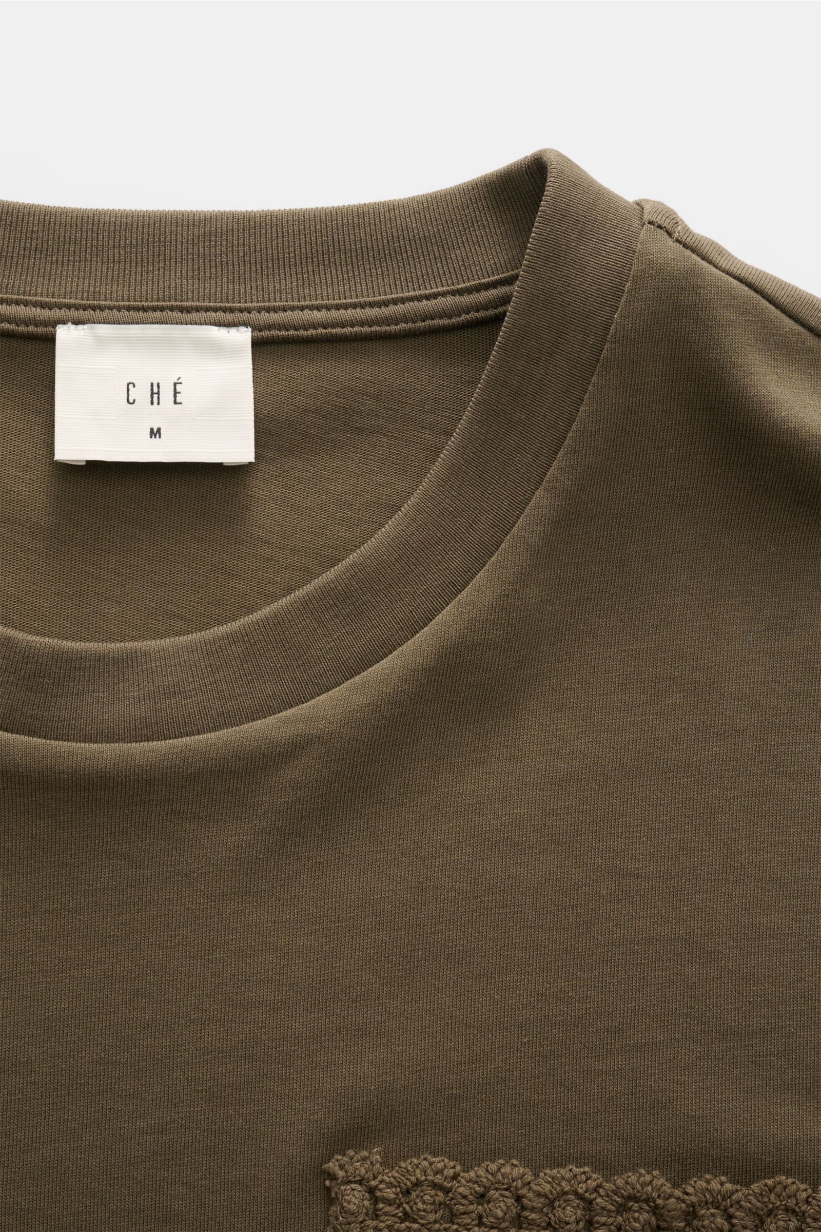 Close-up front top view of Ché T-Shirt 'Inigo' dark olive, showing round neckline, soft cotton fabric, and crochet detail at hem. Sommerliche Lässigkeit: Die Das 'Inigo' T-Shirt von CHÉ wird mit seinen Häkel-Details zum stilvollen Blickfang für entspannte
