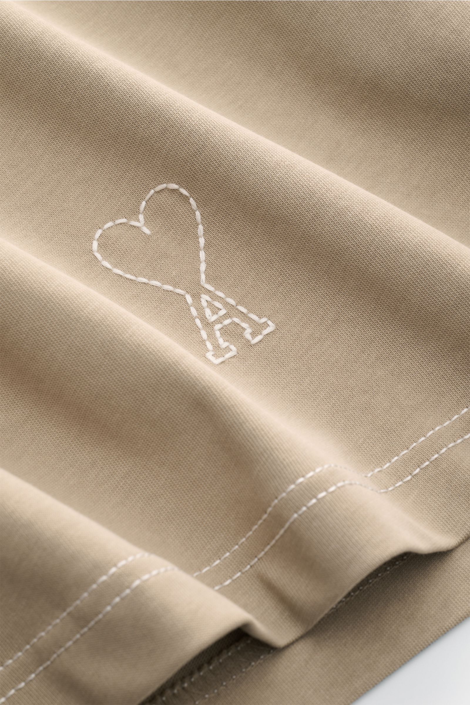 A close-up front view of the Ami Paris Rundhals-T-Shirt taupe, showing the stitched heart logo and double-stitched hem. Mit diesem T-Shirt von AMI PARIS können Sie coole Streetstyles kombinieren - die abgesteppten Nähte und das typische Herz-Logo am Sa