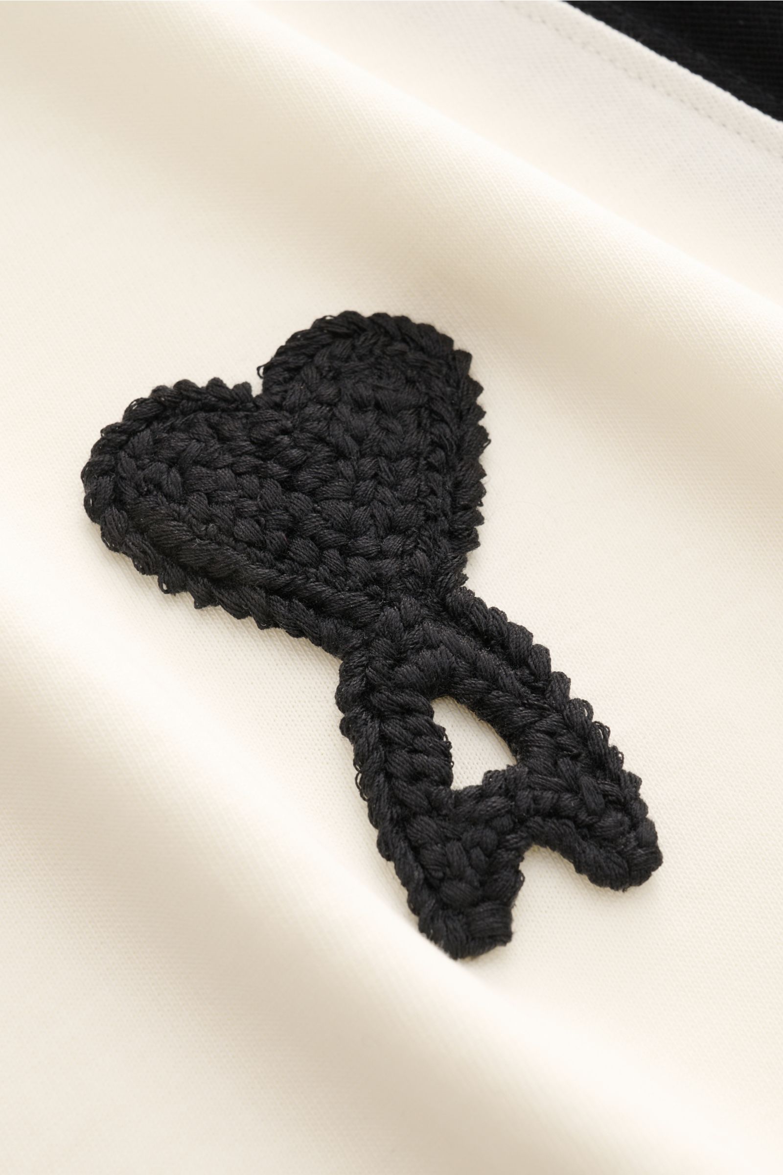 Nahaufnahme des Ami Paris T-Shirts 'Boxy Fit' creme/schwarz mit weichem Baumwollstoff, Raglanärmeln, Rundhalsausschnitt und schwarzem Logo-Stitching auf der Brust, fotografiert von schräg oben.