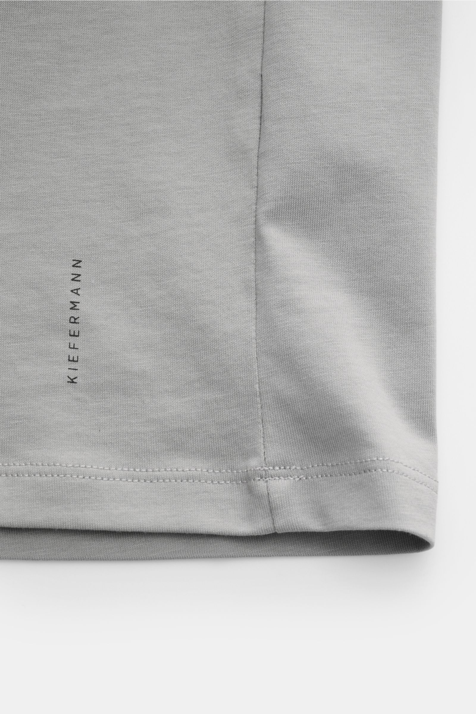 Nahaufnahme des Saums vom Kiefermann Rundhals T-Shirt 'Matello' grau, extra slim Fit, Baumwoll-Jersey mit weichem, glattem Griff, Rundhalsausschnitt, dezenter Logodruck am Saum, von vorne fotografiert.