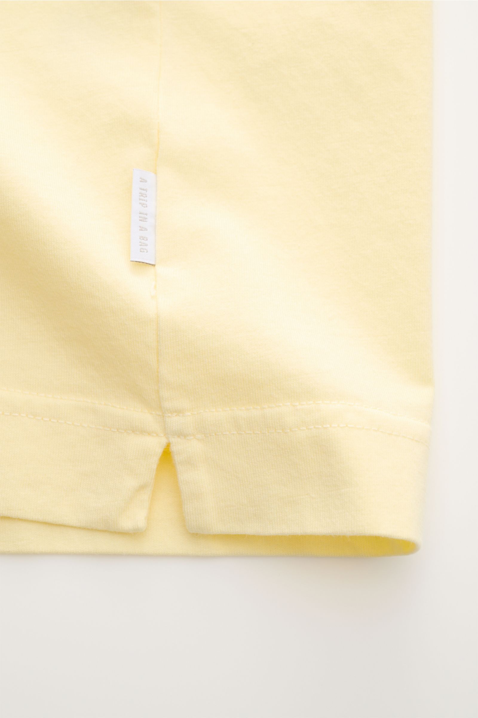 Nahaufnahme des Saumabschnitts vom 04651/ A trip in a bag Rundhals-T-Shirt pastellgelb, Jersey aus reiner Baumwolle, weicher Griff, Regular Fit, Rundhalsausschnitt, kleines Weblabel sichtbar.