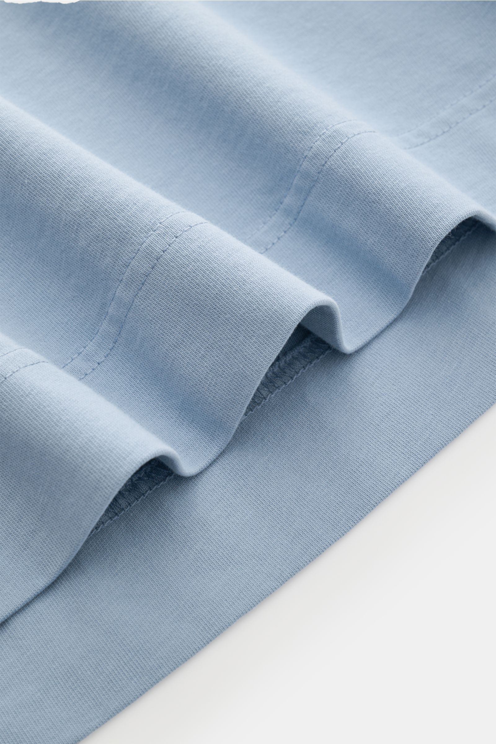 Close-up from a side angle of the hem detail of the 04651/ A trip in a bag T-shirt 'Nunu' smoky blue, showing soft pure cotton fabric with neat stitching. A basic with a print accent: With its minimalist holiday print, this T-shirt by 04651/ A TRIP IN A B