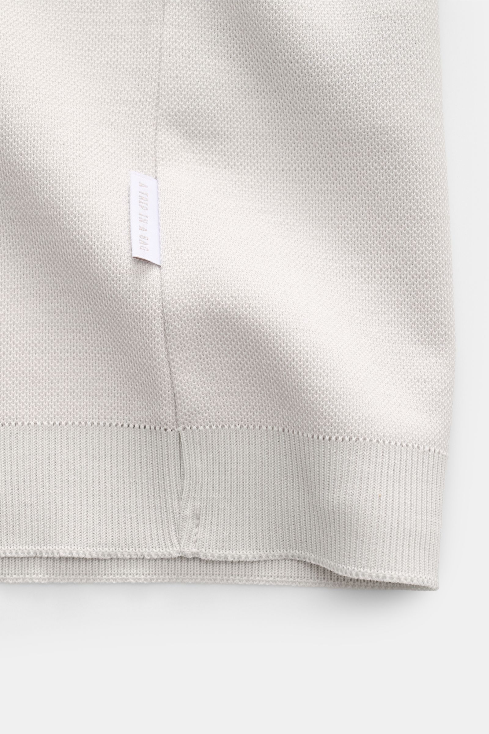 Nahaufnahme des hellgrauen 04651/ A trip in a bag Piqué-T-Shirts 'TP' mit sichtbarem Weblabel an der Seitennaht, Piqué-Struktur und Rippbündchen, Slim Fit, Rundhalsausschnitt, aus reiner Baumwolle mit kernigem Griff, fotografiert aus schräger Perspektive.