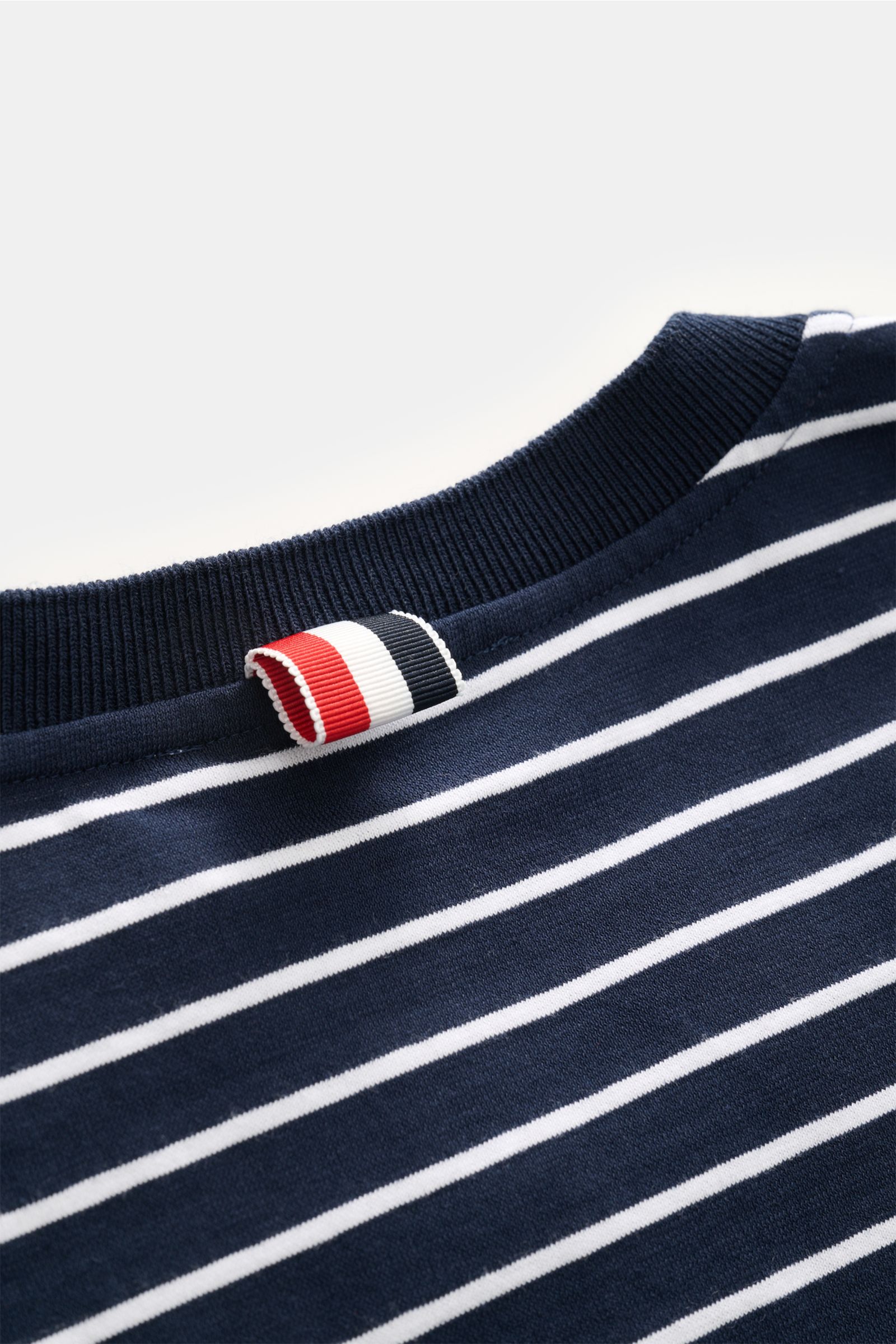 Close-up view of the neckline of the Thom Browne Rundhals-T-Shirt navy/weiß gestreift, showing navy blue ribbed collar, white horizontal stripes, and brand's signature red, white, and navy tab.

Zeitlos maritim: Das T-Shirt von THOM BROWNE aus reiner B