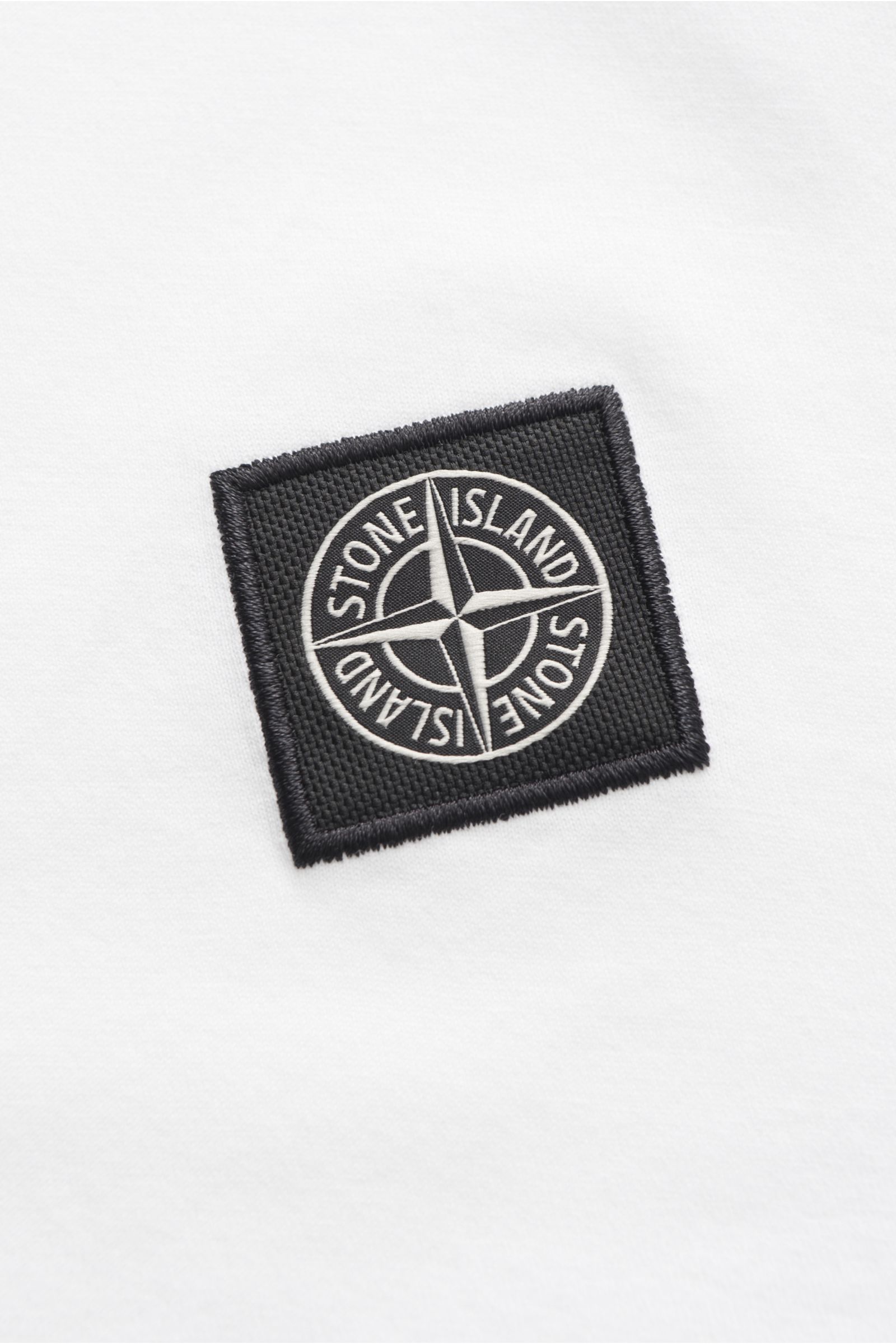 Stone Island Rundhals-Longsleeve weiß, Detailfoto des schwarzen Kompass-Logos auf weichem, weißem Baumwolljersey mit Rippbündchen.