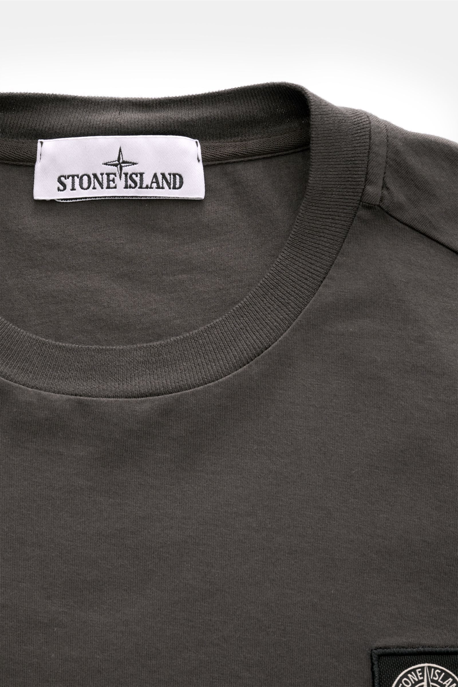 Close-up front top view of Stone Island Rundhals-Longsleeve 'Basic Cotton' dunkelgrau, a slim fit dark gray long sleeve with round neckline, soft cotton fabric, ribbed cuffs, and a Stone Island web label on the chest. Zeitloses Basic Piece: Das Longsle