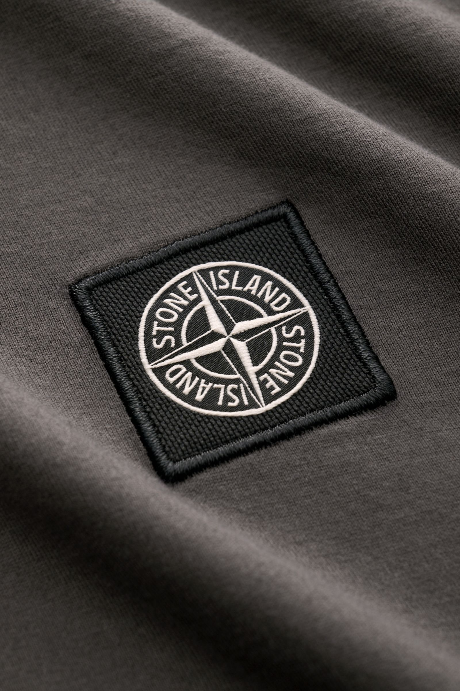 Close-up front view of Stone Island Rundhals-Longsleeve 'Basic Cotton' dunkelgrau showing black embroidered logo patch on dark gray cotton fabric.

Zeitloses Basic Piece: Das Longsleeve von STONE ISLAND rundet legere Freizeitlooks mühelos ab und wird z