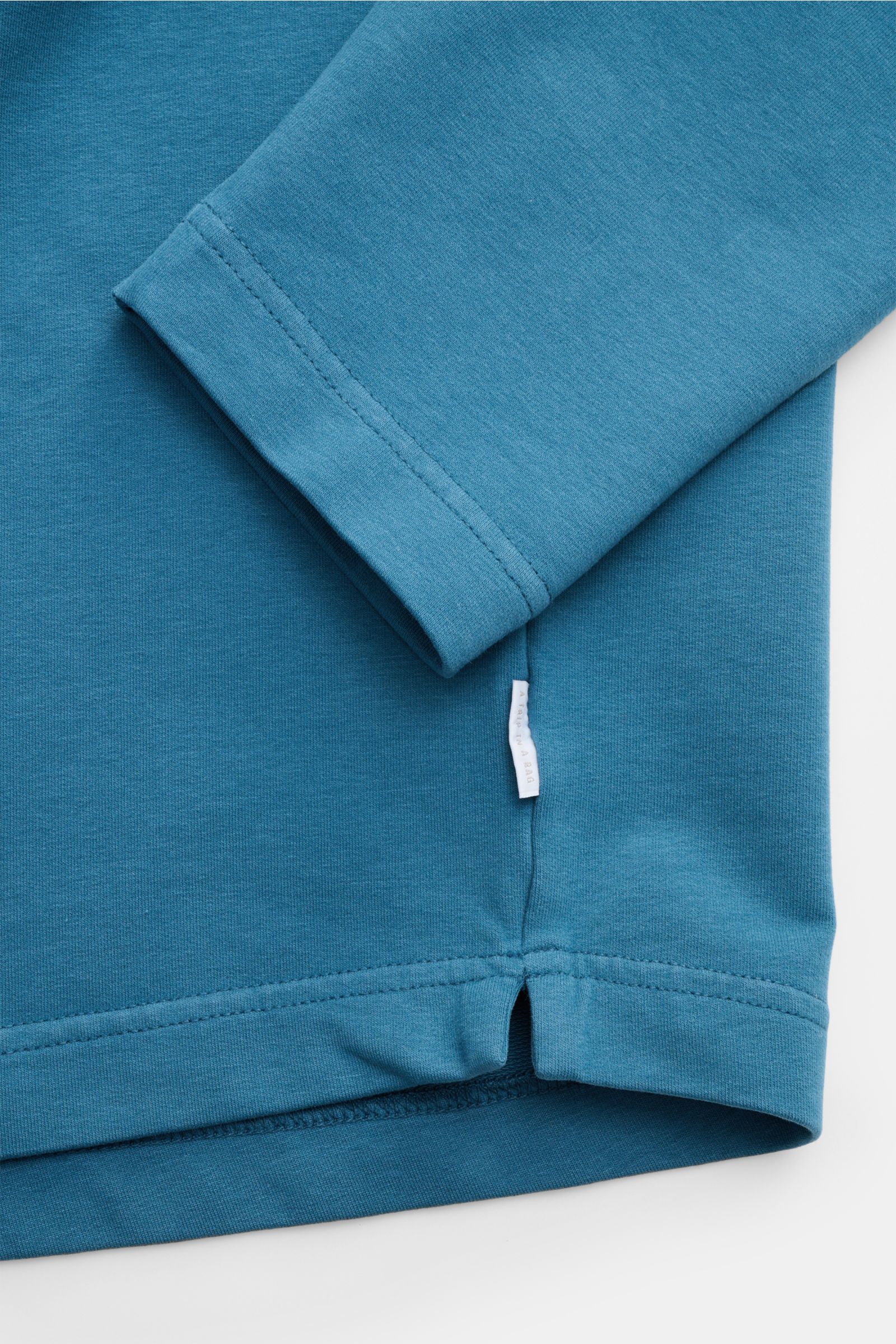 Nahaufnahme von Ärmel und Saum des petrolfarbenen 04651/ A trip in a bag Rundhals-Longsleeve 'If Tee', Jersey aus elastischer Baumwolle, Oversized Fit, Weblabel seitlich.