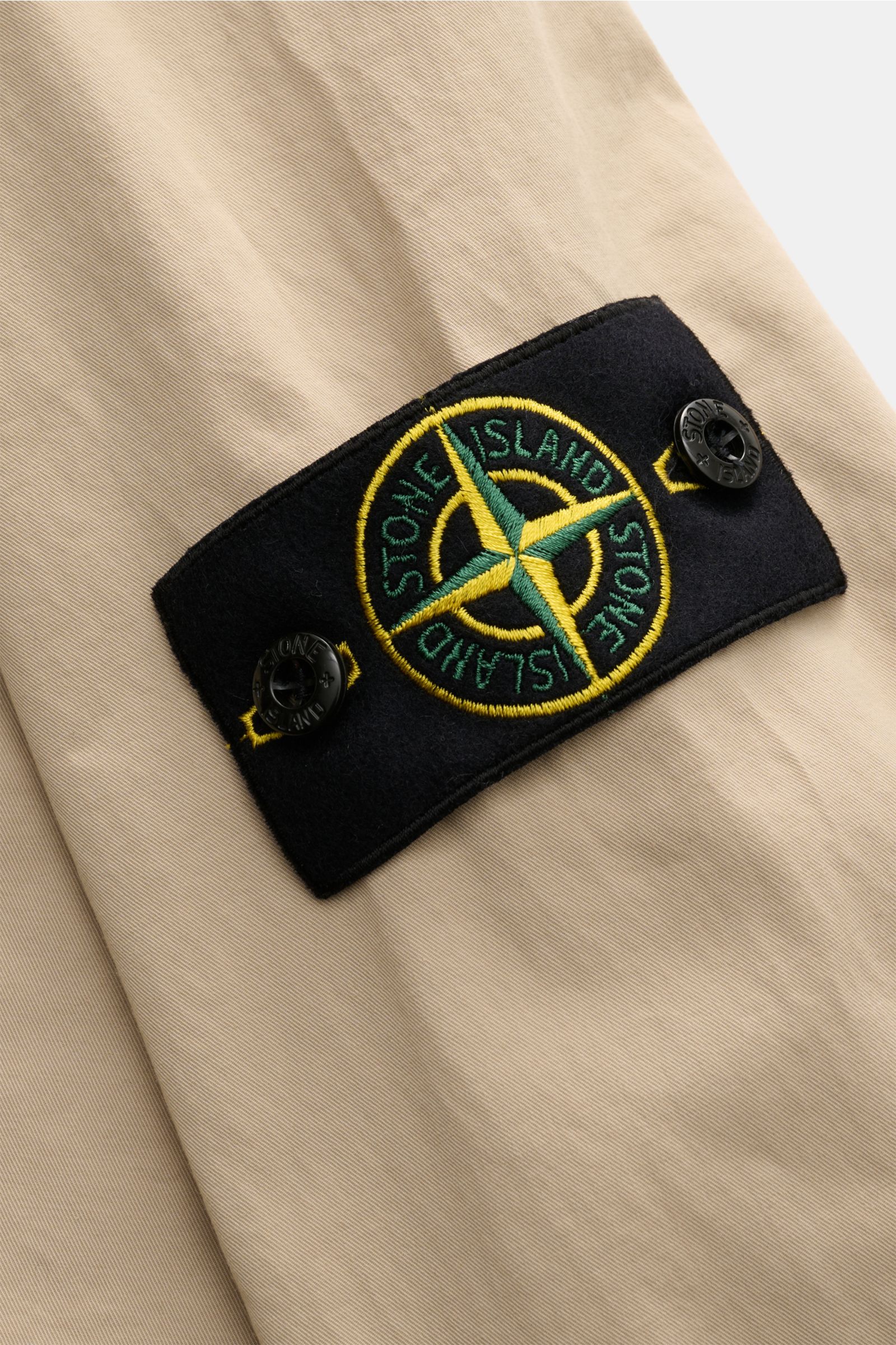 Nahaufnahme des Kompass-Badges am Ärmel des Stone Island Overshirt beige aus Twill, Baumwolle, elastisch, weich, leicht, garment dyed, Slim Fit, mit zwei-Wege-Reißverschluss, Umlegekragen, Brusttasche mit Zipper und regulierbaren Ärmelabschlüssen.