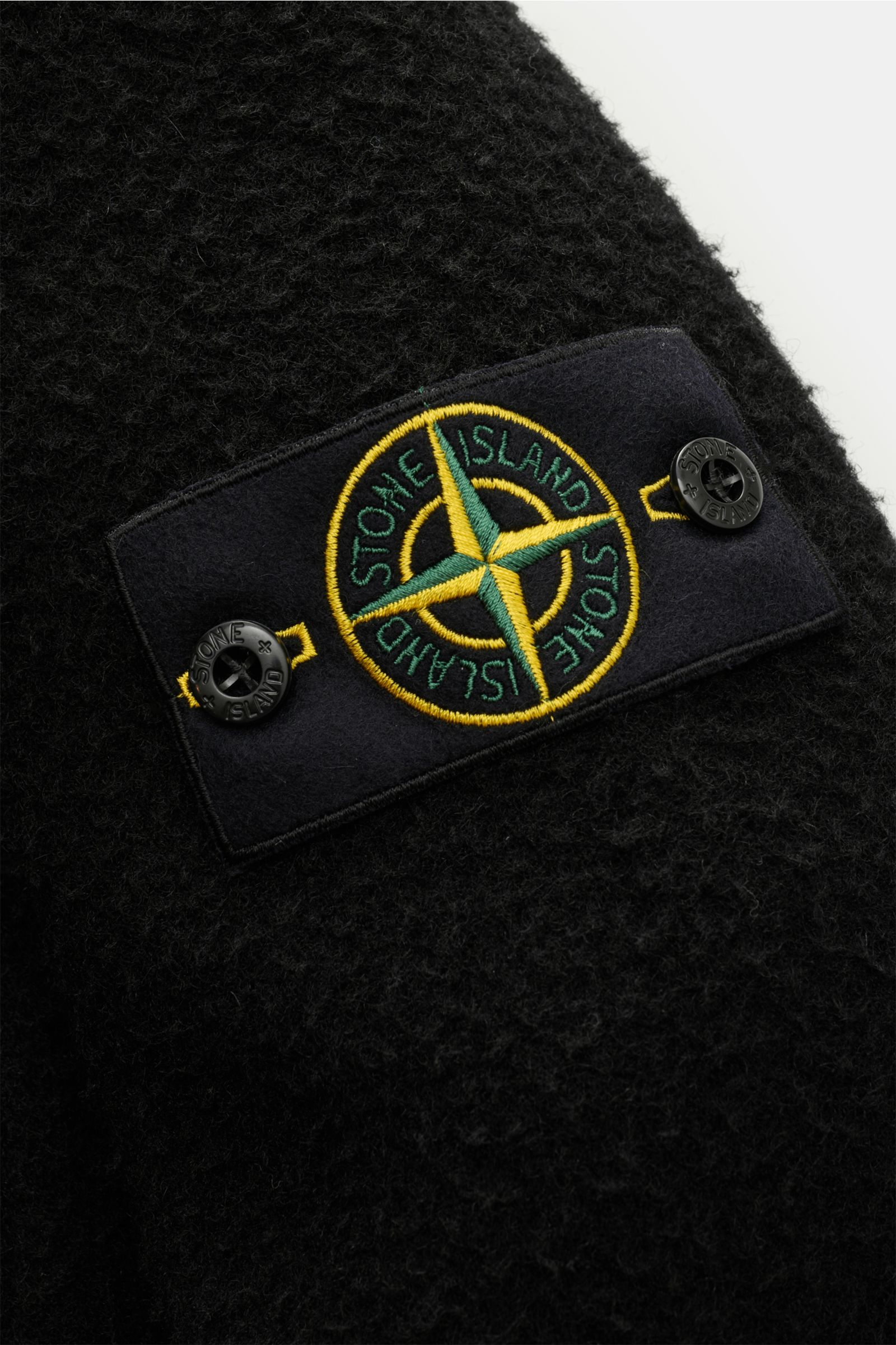 Nahaufnahme der schwarzen Stone Island Jacke 'Panno Ratinato' von schräg oben, zeigt Kompass-Badge am Ärmel. Mix aus Wolle und Microfaser, strukturierte Haptik, Slim Fit, Druckknopfleiste, Umlegekragen, Ärmelabschlüsse mit Druckknopf, regulierbare Saumwei