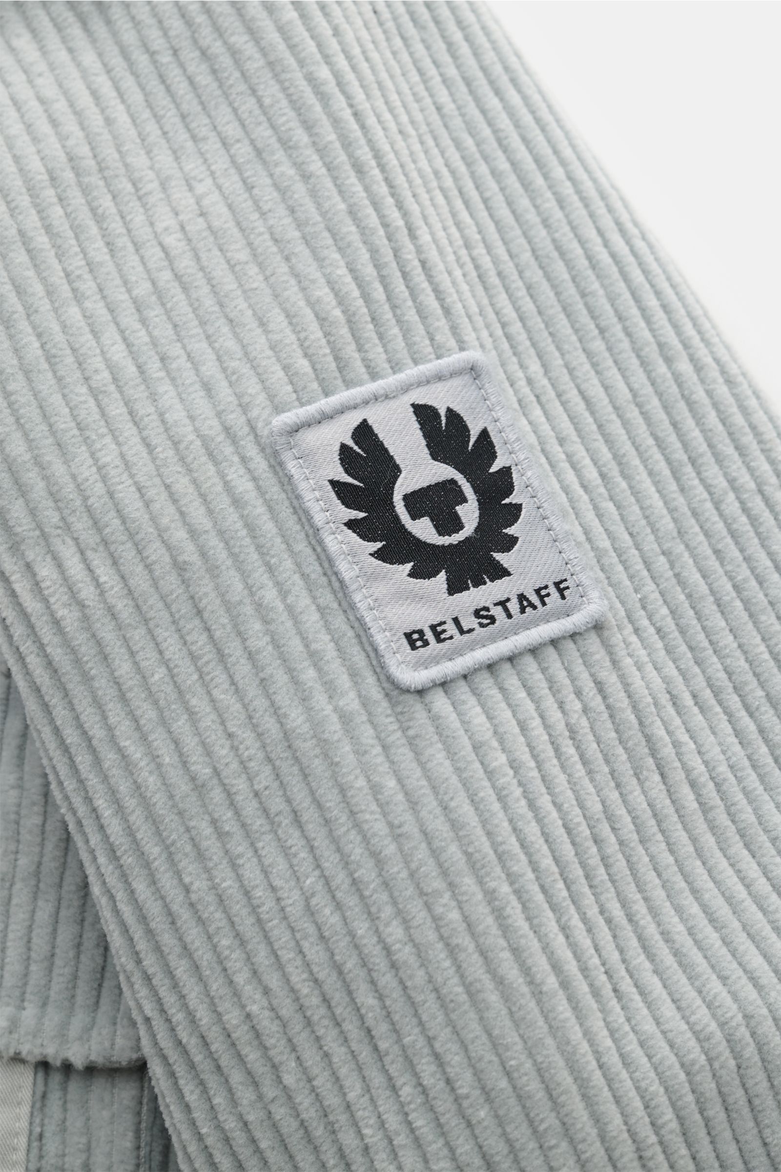 Nahaufnahme des tonalen BELSTAFF-Logos am Ärmel des Belstaff Cord-Overshirt 'Fallgate' grau aus robustem Baumwollcord, slim fit, mit Twill-Einsätzen.