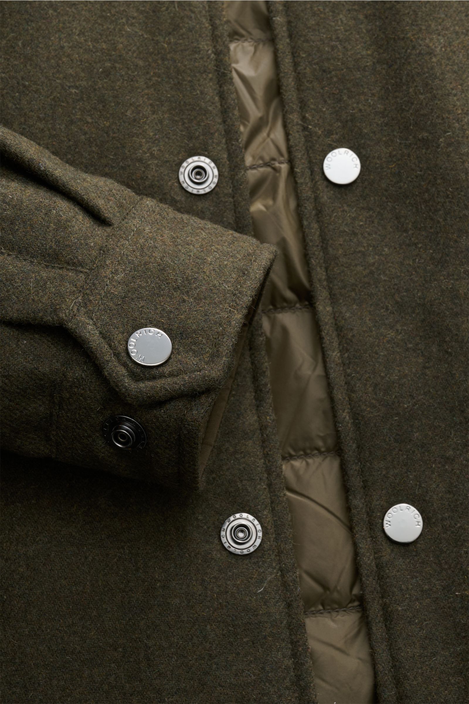 Nahaufnahme der Woolrich Daunenjacke dark olive aus der Vogelperspektive, zeigt Mix aus Wolle und Microfaser, leichte Daunenfüllung, Slim Fit, Druckknopfleiste, variablen Kragen mit Knopfriegel, schräge Leistentaschen, Ärmelabschluss mit Knopf, regulierba