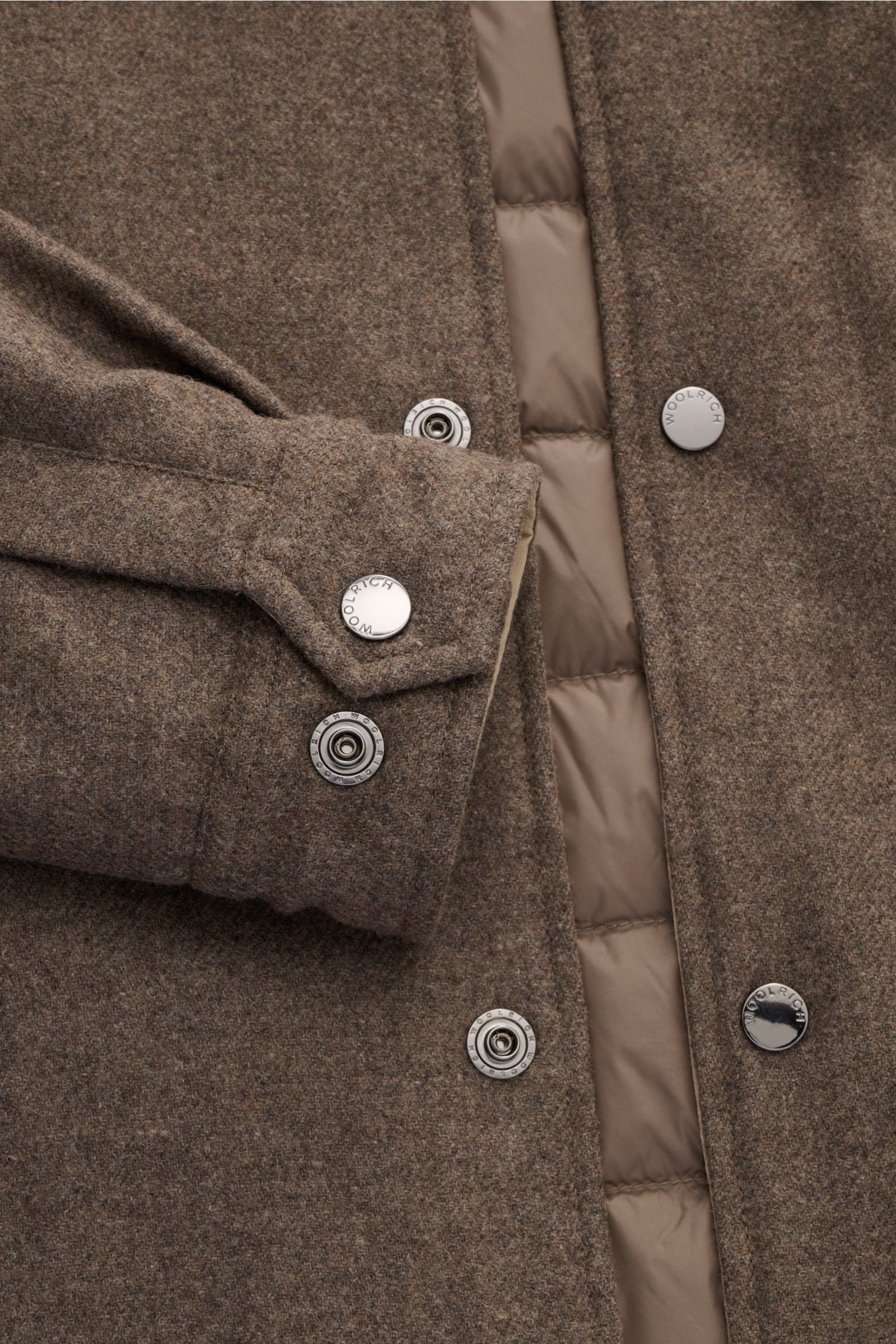 Nahaufnahme der Woolrich Daunenjacke taupe von oben, zeigt Wolle und Microfaser, leichte Daunenfüllung, Slim Fit, Druckknopfleiste, variablen Kragen mit Knopfriegel, schräge Leistentaschen, Ärmelabschlüsse mit Knopf, regulierbare Taillenweite, gesteppten,