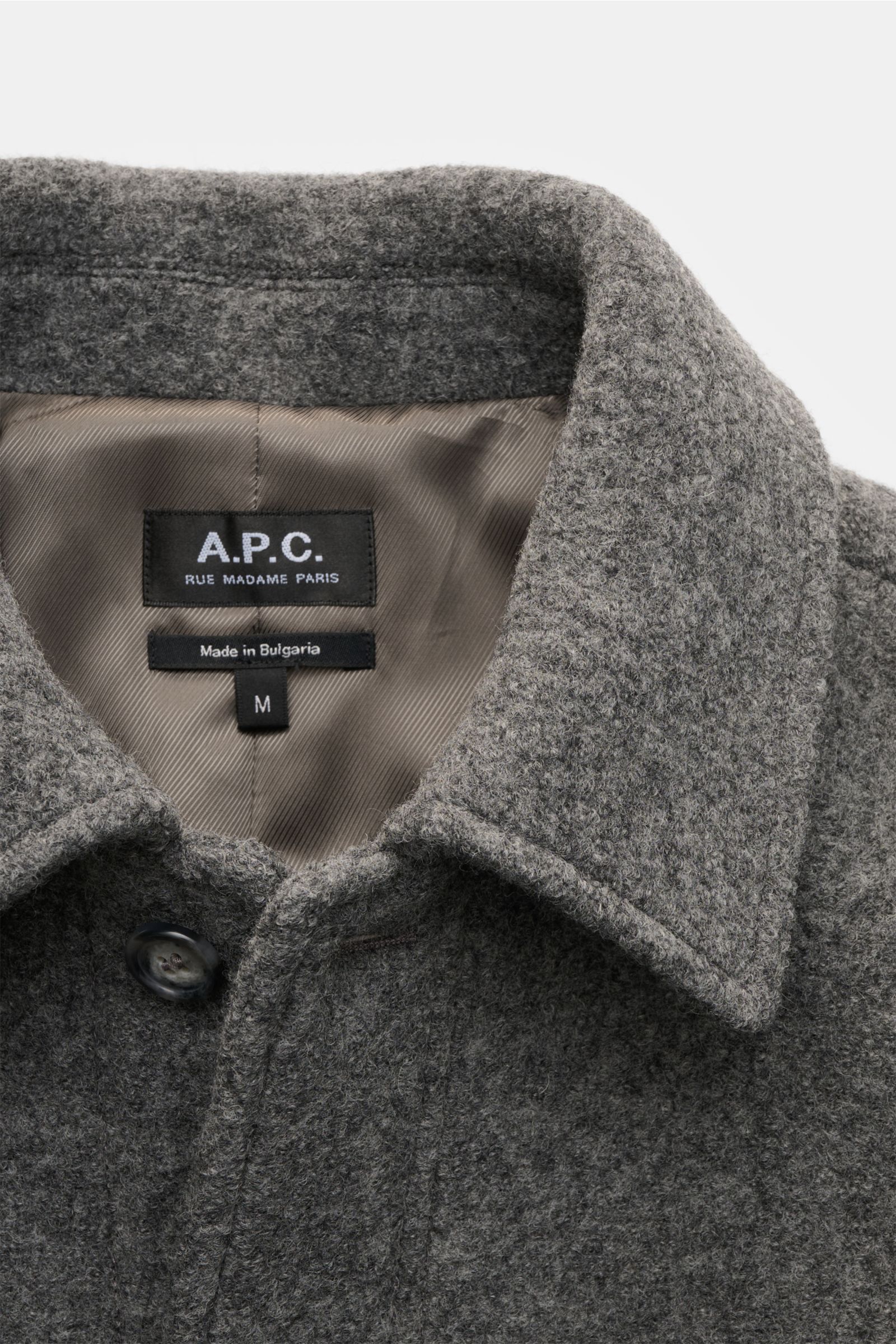 A close-up front view of the A.P.C. Bouclé-Overshirt 'Emile' dunkelgrau showing its grey wool bouclé fabric, folded collar, button, and inner tonal lining with label. Anziehen, wohlfühlen, cool aussehen: Das Overshirt 'Emile' von A.P.C. aus Woll-Bouclé