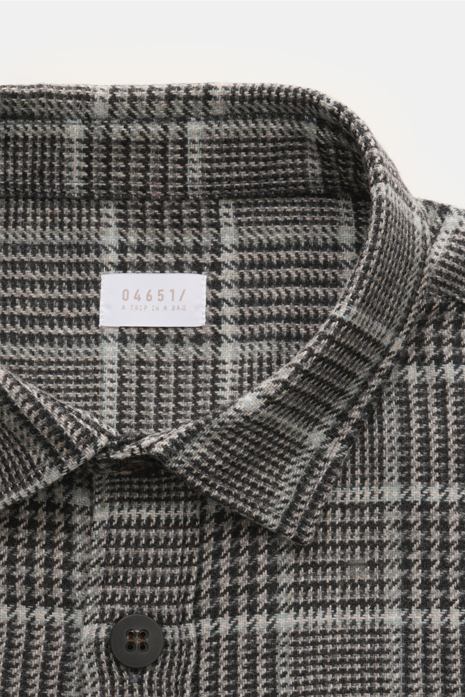 Close-up front view of the 04651/ A trip in a bag Overshirt schwarz/grau kariert showing the collar, top button, and Glencheck pattern.  
Das perfekte Styling-Piece für Ihre Freizeitgarderobe: Das karierte Overshirt von 04651/ A TRIP IN A BAG wurde aus