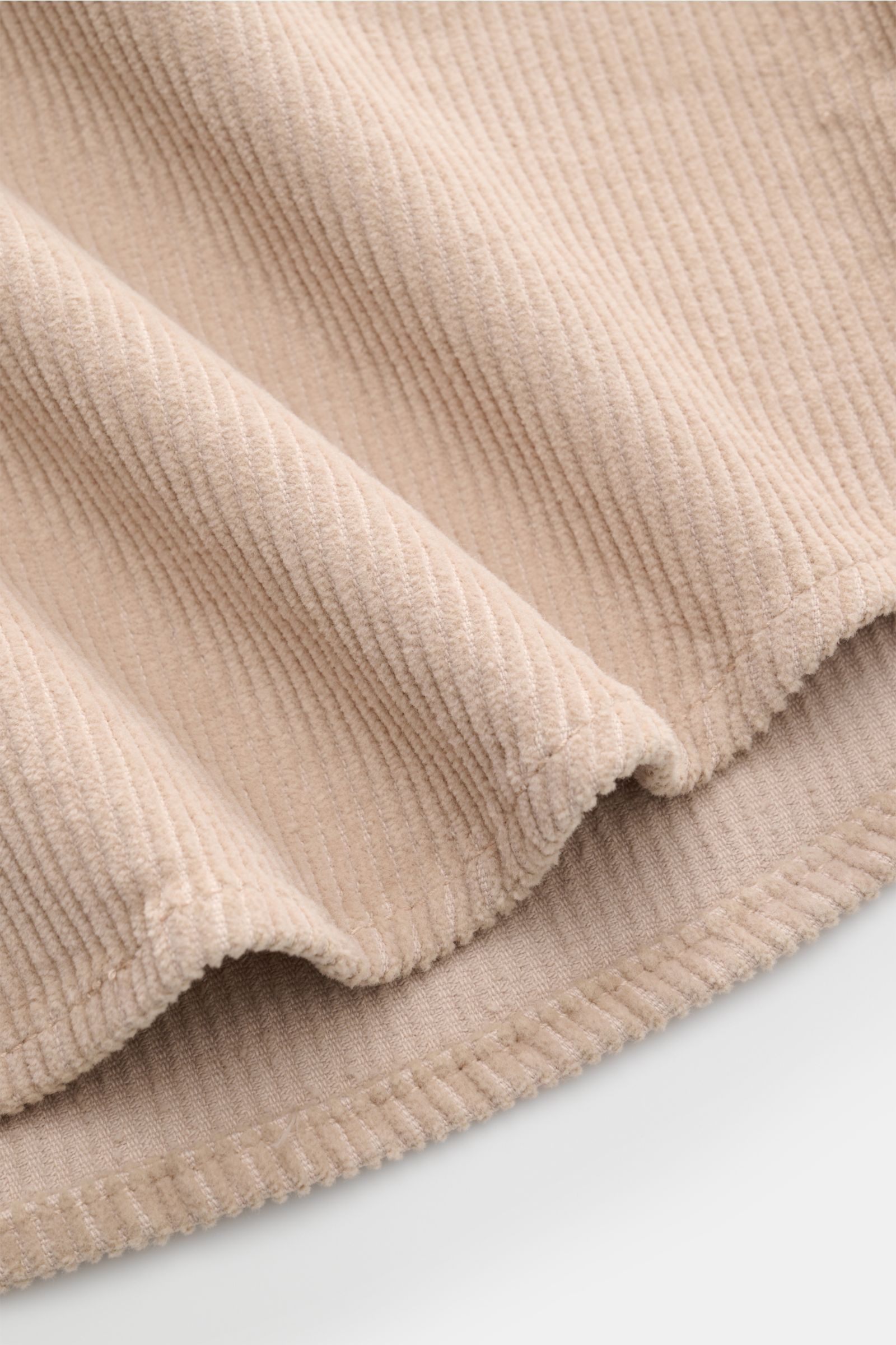 Close-up of the beige Ace Denim Cord-Overshirt fabric showing its soft pure cotton corduroy texture, photographed from a side angle highlighting the rounded hem and fine stitching.