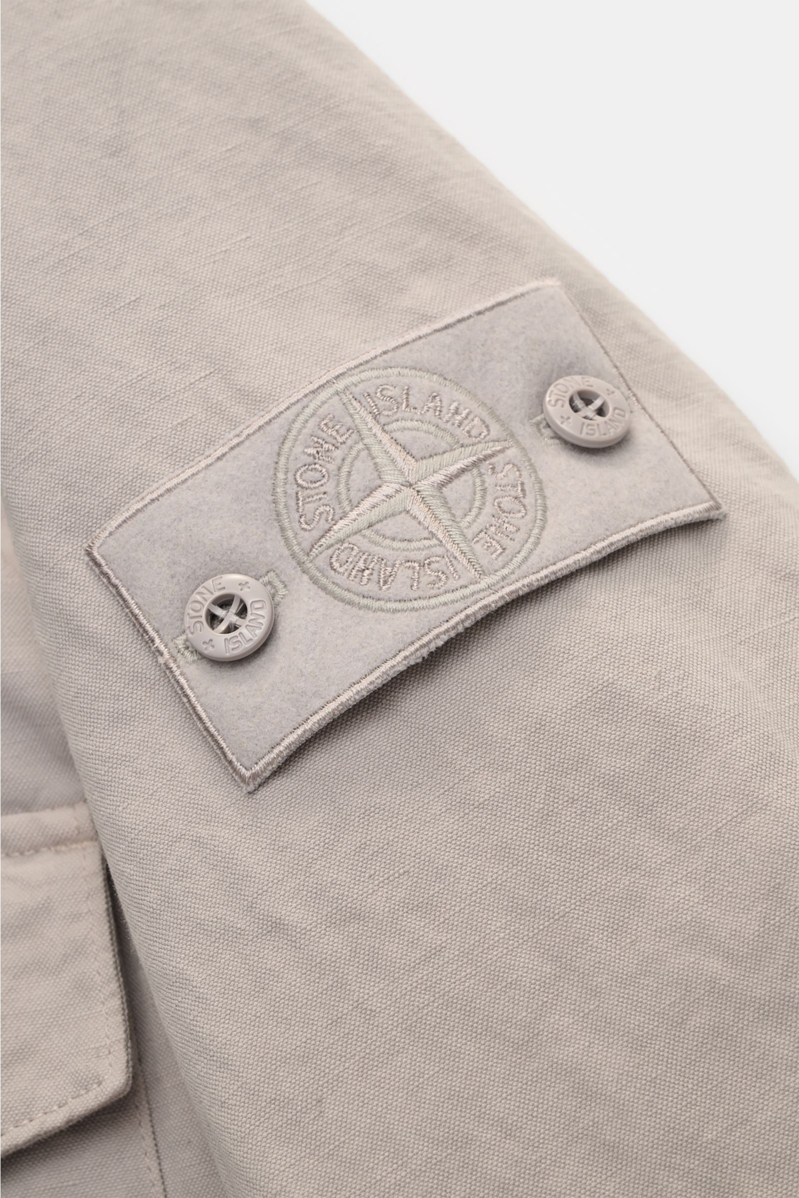 Close-up side view of the compass badge on the sleeve of the Stone Island Overshirt 'Ghost' light grey, a timeless layering piece with a cotton and linen blend, slim fit, six-button placket, turn-down collar, three front pockets, cuffs with button, and un