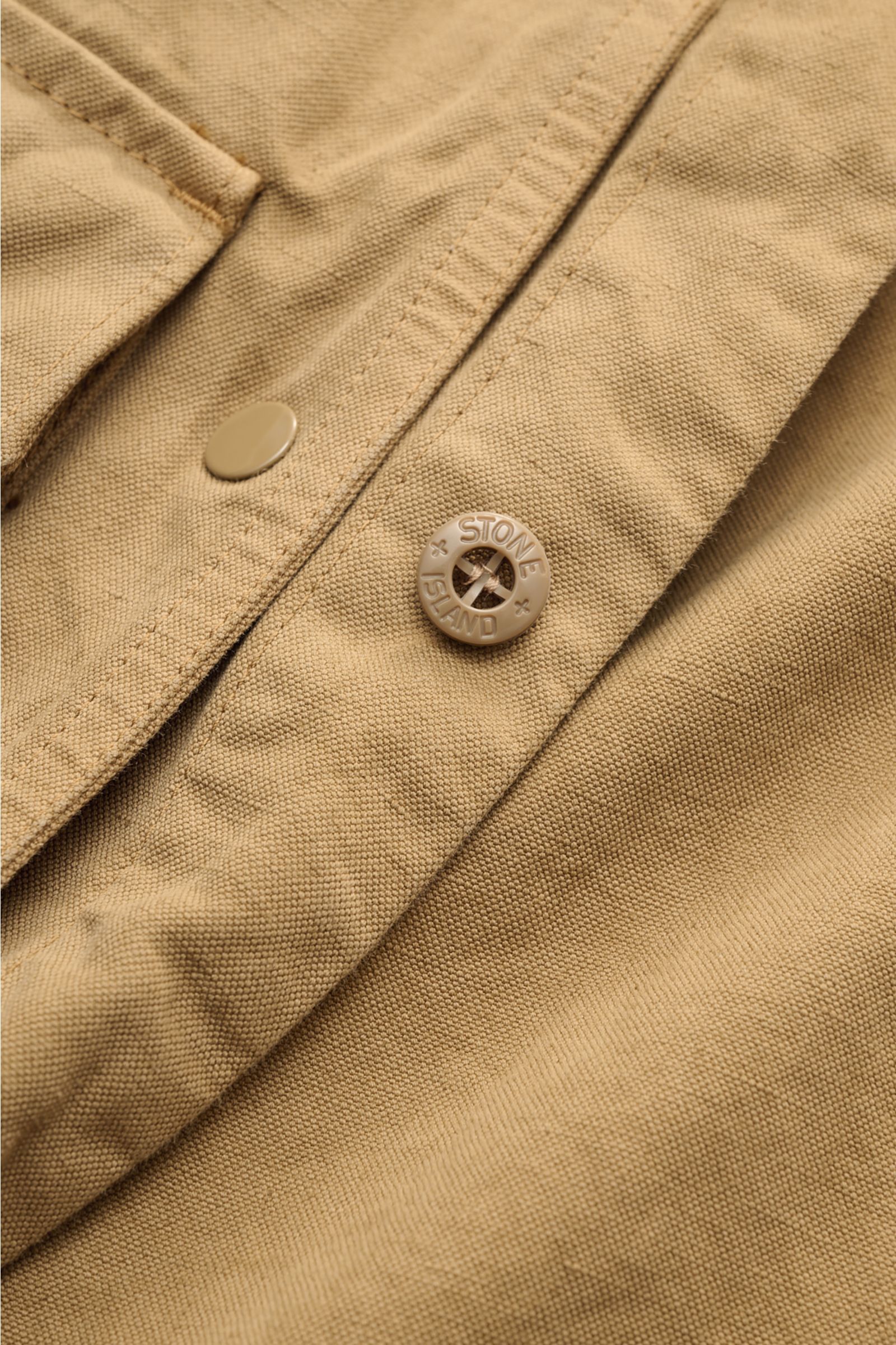 Stone Island Overshirt 'Ghost' khaki close-up showing textured cotton-linen fabric, button detail, and stitching from above angle.  
Zeitloses Layering-Piece: Das Overshirt aus der 'Ghost' Capsule von STONE ISLAND ergänzt lässige Frühlingslooks perfekt