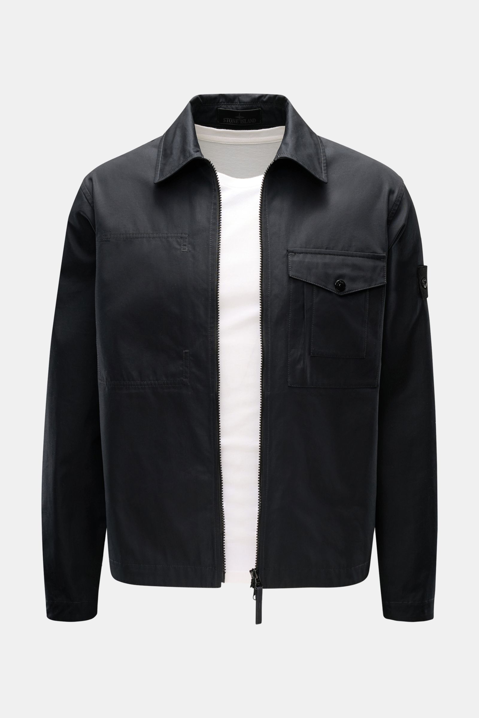Front view of Stone Island Overshirt 'Ghost Piece' black, a slim-fit black cotton canvas jacket with turn-down collar, two-way zip, breast pocket with flap, tonal sleeve badge, and button cuffs, worn over a white shirt. Function meets comfort: This STONE 