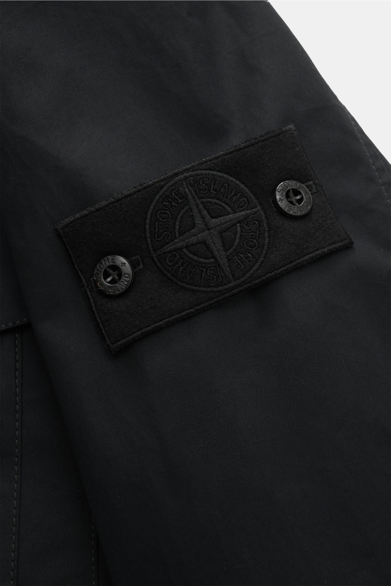 Close-up side view of the tonal black Stone Island Overshirt 'Ghost Piece' black showing the embroidered badge on the upper sleeve. Function meets comfort: This STONE ISLAND overshirt from the 'Ghost' capsule is made of uniquely comfortable cotton canvas 