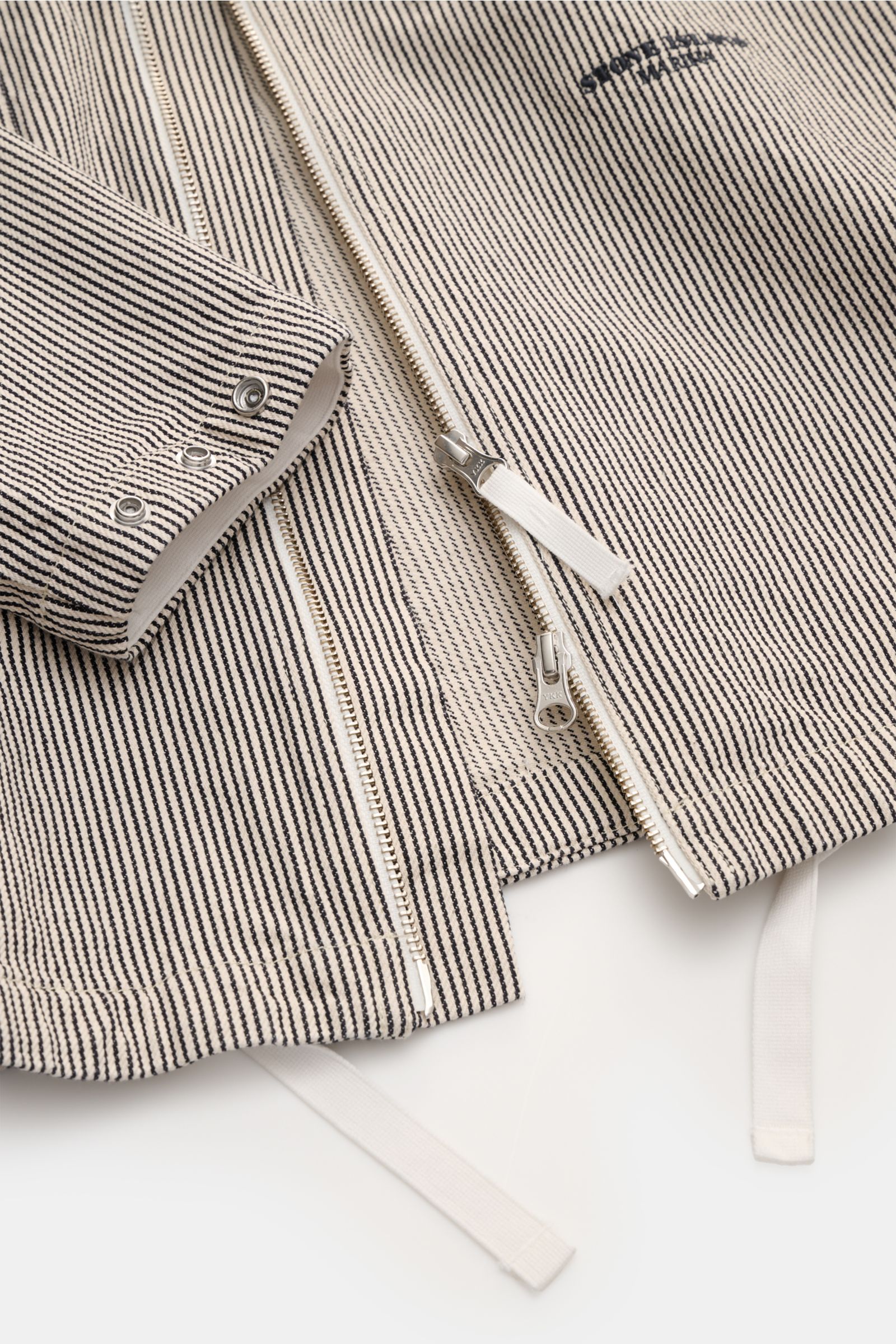 Close-up top-down view of the Stone Island Jacke 'Marina' navy/beige gestreift showing the striped pure cotton fabric, robust two-way zipper, snap button cuff, adjustable hem, front chest pocket, and Marina stitching. Leichte Outerwear mit maritimem Chara