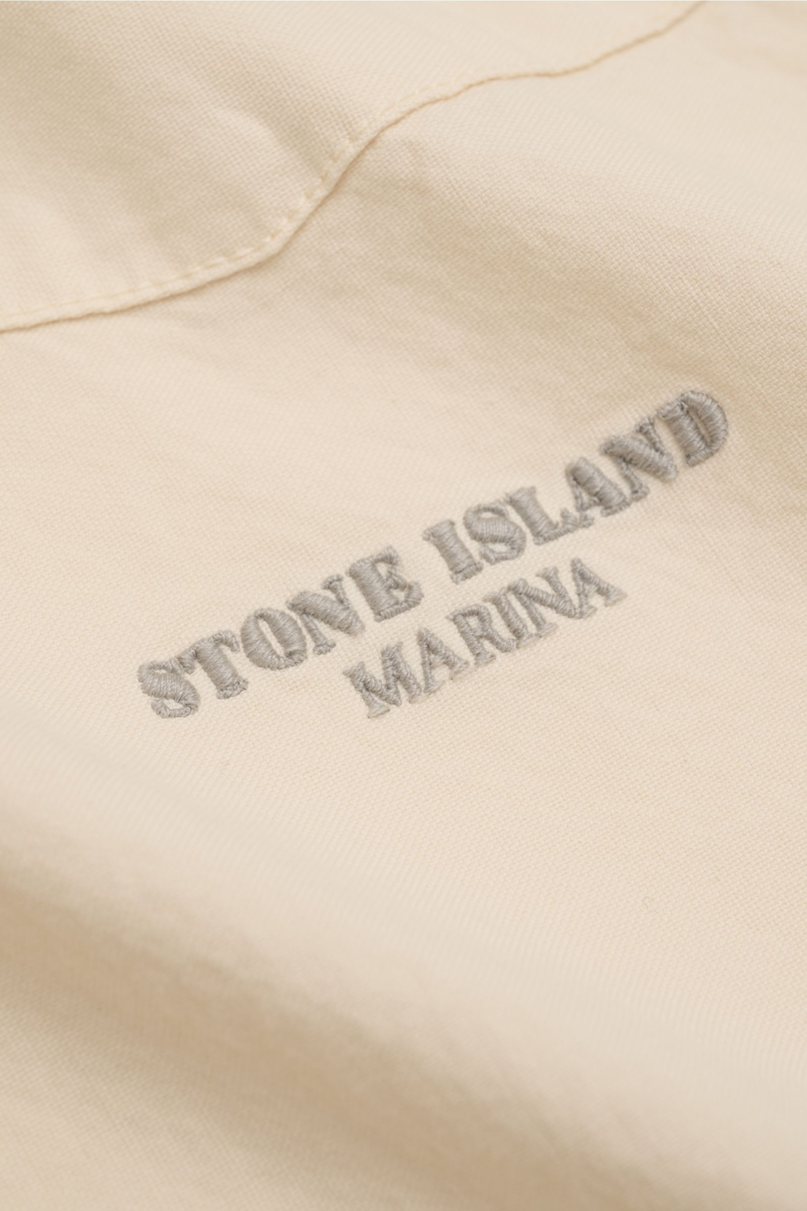 Close-up view of the Stone Island Overshirt 'Marina' creme showing detailed gray MARINA stitching on soft, creamy Panama cotton fabric. Maritimes Layering-Piece: Das Overshirt aus der STONE ISLAND 'Marina' Capsule Collection verbindet leichtes Panama-Gewe