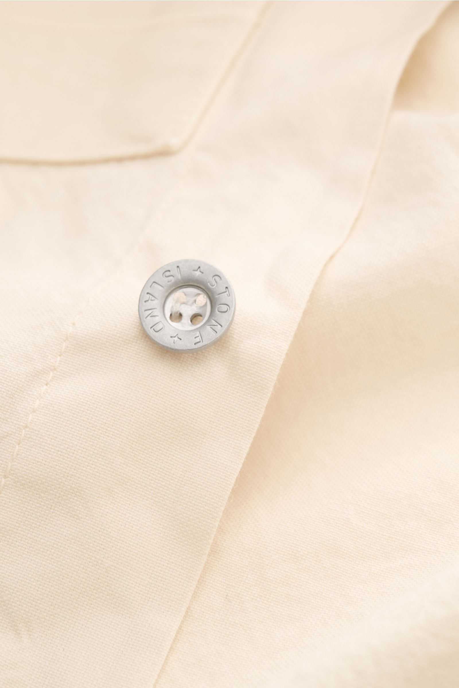 Close-up front view of the Stone Island Overshirt 'Marina' creme showing a silver branded button on soft, cream-colored pure cotton Panama weave fabric with a smooth texture. Maritimes Layering-Piece: Das Overshirt aus der STONE ISLAND 'Marina' Capsule Co