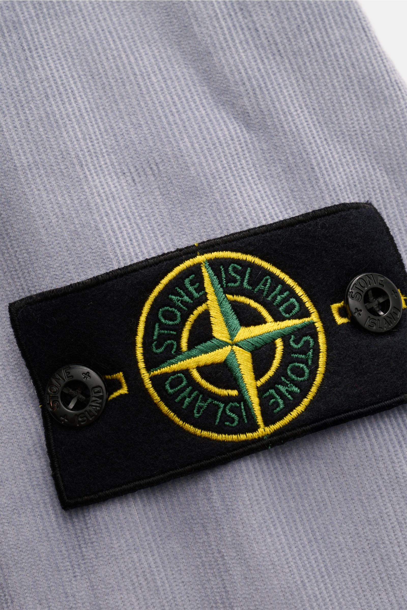 Nahaufnahme des typischen Kompass-Badges am Ärmel des Stone Island Cord Overshirt rauchblau aus Bio-Baumwolle mit weichem Griff.