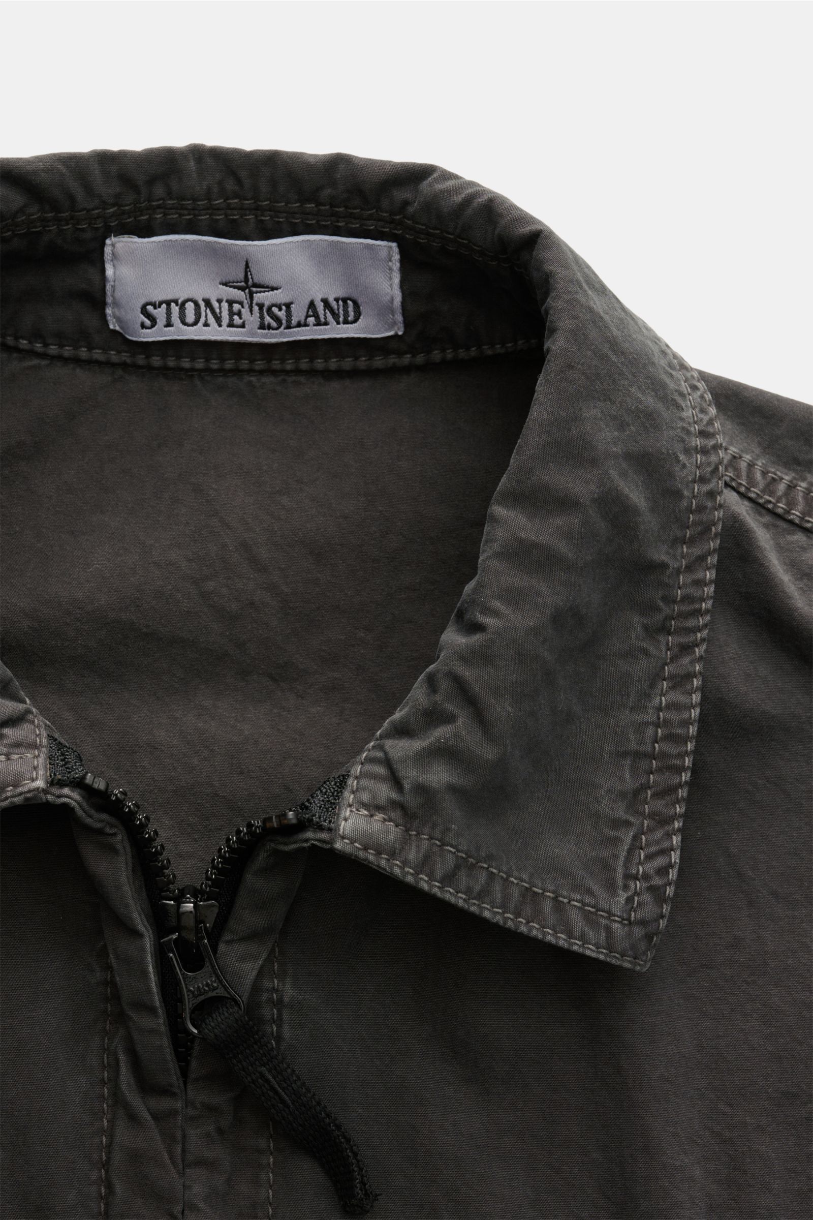 Close-up front view of the Stone Island Overshirt 'Brushed Canvas' dunkelgrau showing the collar, two-way zipper, and brand label inside. Lässiges Layering-Piece: Das Canvas-Overshirt von STONE ISLAND lässt sich mühelos zu legeren Looks kombinieren. Reine