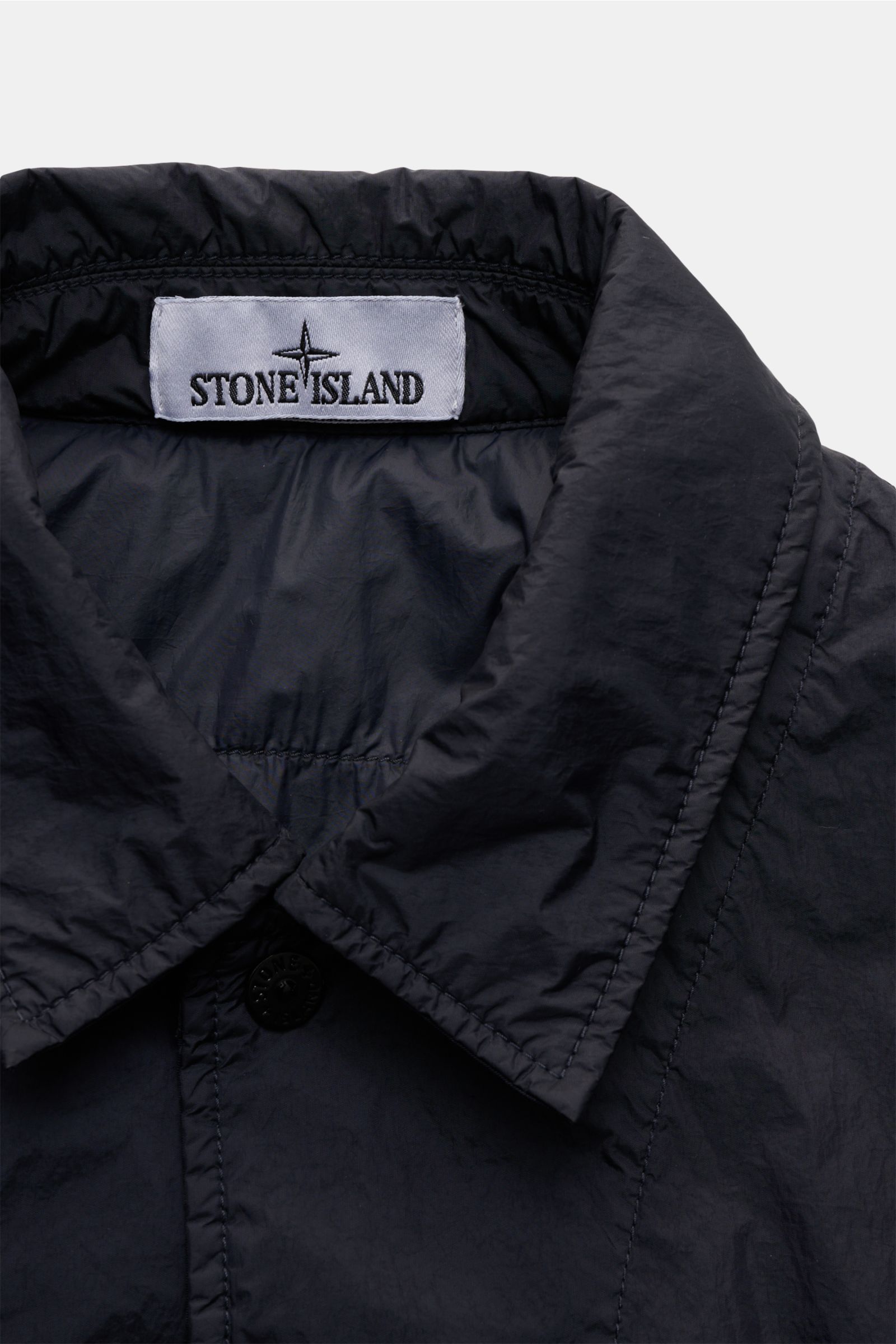 Nahaufnahme des Kragens vom Stone Island Overshirt 'Crinkle Reps' navy, wind- und wasserabweisend, garment dyed, mit Druckknopfleiste und Kompass-Badge.
