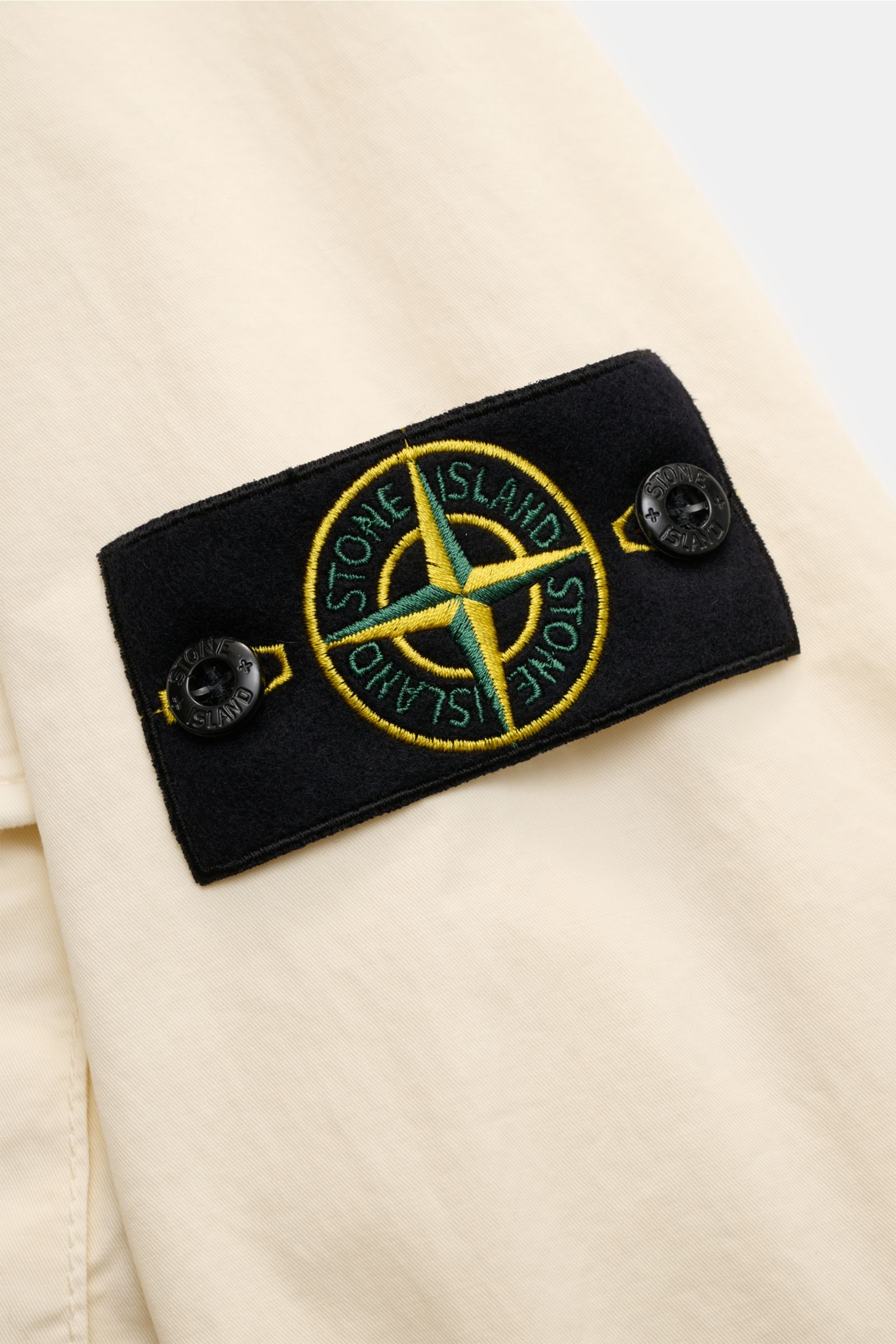 Close-up side view of Stone Island Overshirt 'Stretch Cotton' creme, showing the black compass badge with yellow and green embroidery on the cream fabric.

Description: Sportives Overshirt mit urbaner Lässigkeit: Dieser markante Hybrid aus Casual Hemd 
