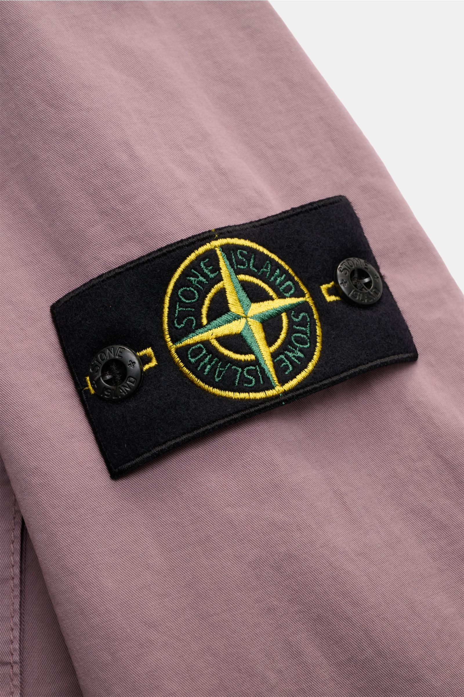 Close-up from the side of the Stone Island Overshirt 'Stretch Cotton' flieder showing the black compass badge with yellow and green embroidery on soft, garment-dyed lilac cotton fabric.

Description: Sportives Overshirt mit urbaner Lässigkeit: Dieser m