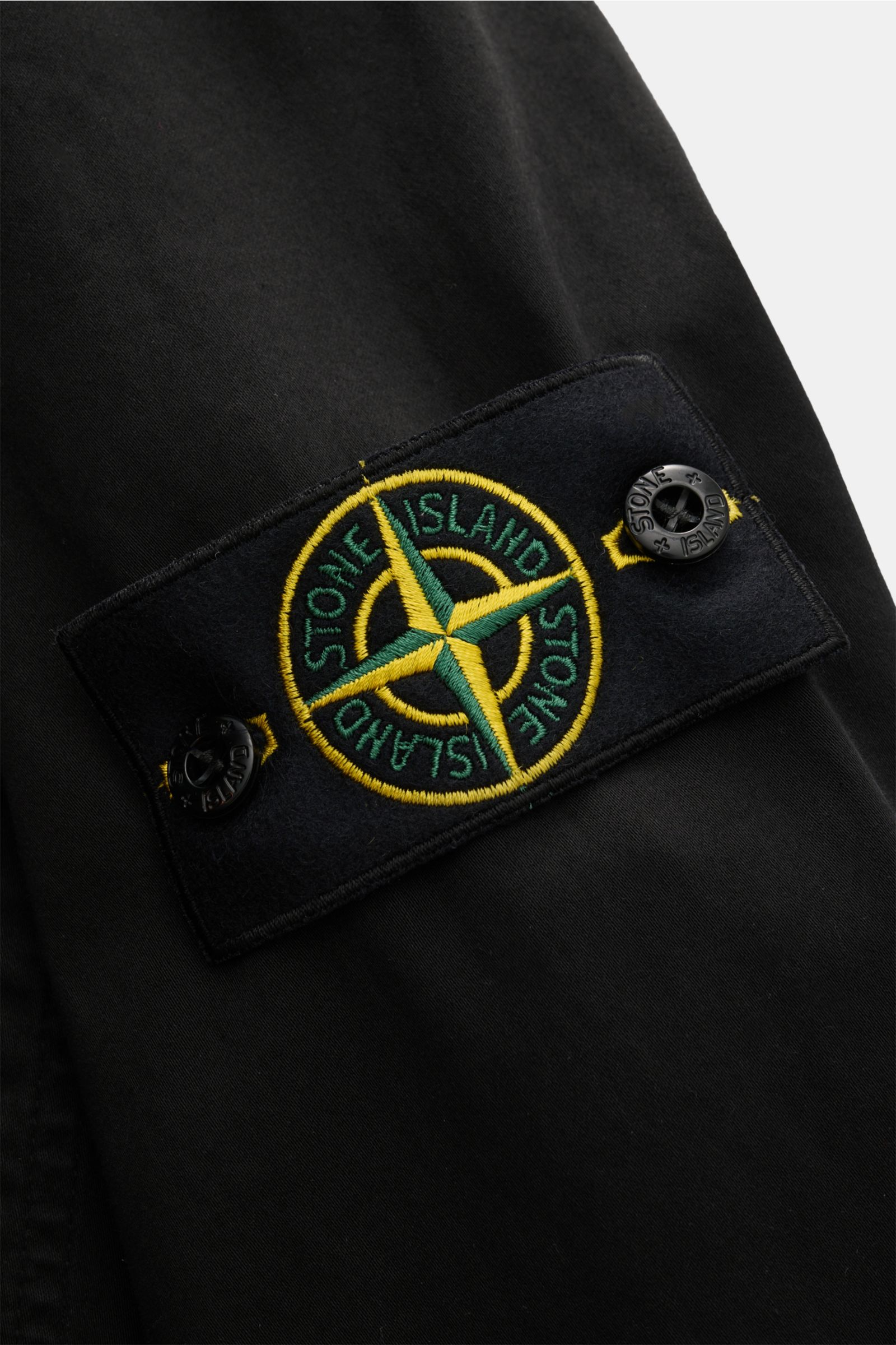 Nahaufnahme des Kompass-Badges am Ärmel des Stone Island Overshirt 'Cotton Satin' schwarz aus elastischer Baumwolle mit Knopfverschluss.