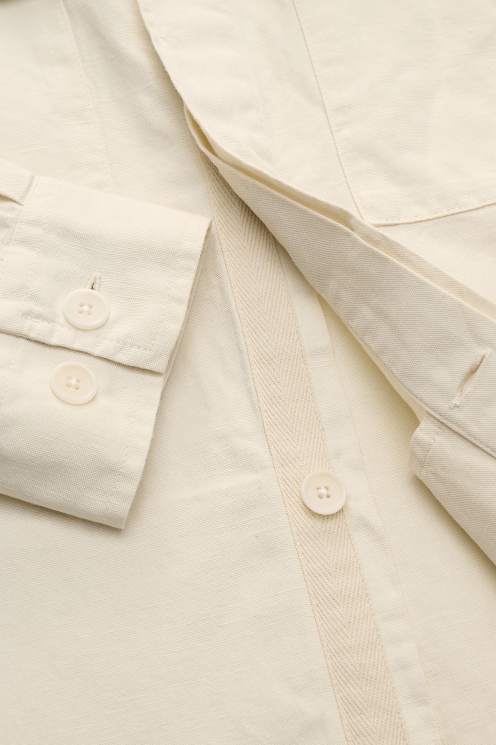 Close-up front detail of Belstaff Overshirt 'Maritime' sand, showing slim fit design, concealed button placket, narrow collar, patch breast pocket with flap, classic barrel cuffs, and woven label on sleeve, photographed from a top-down perspective in a ro