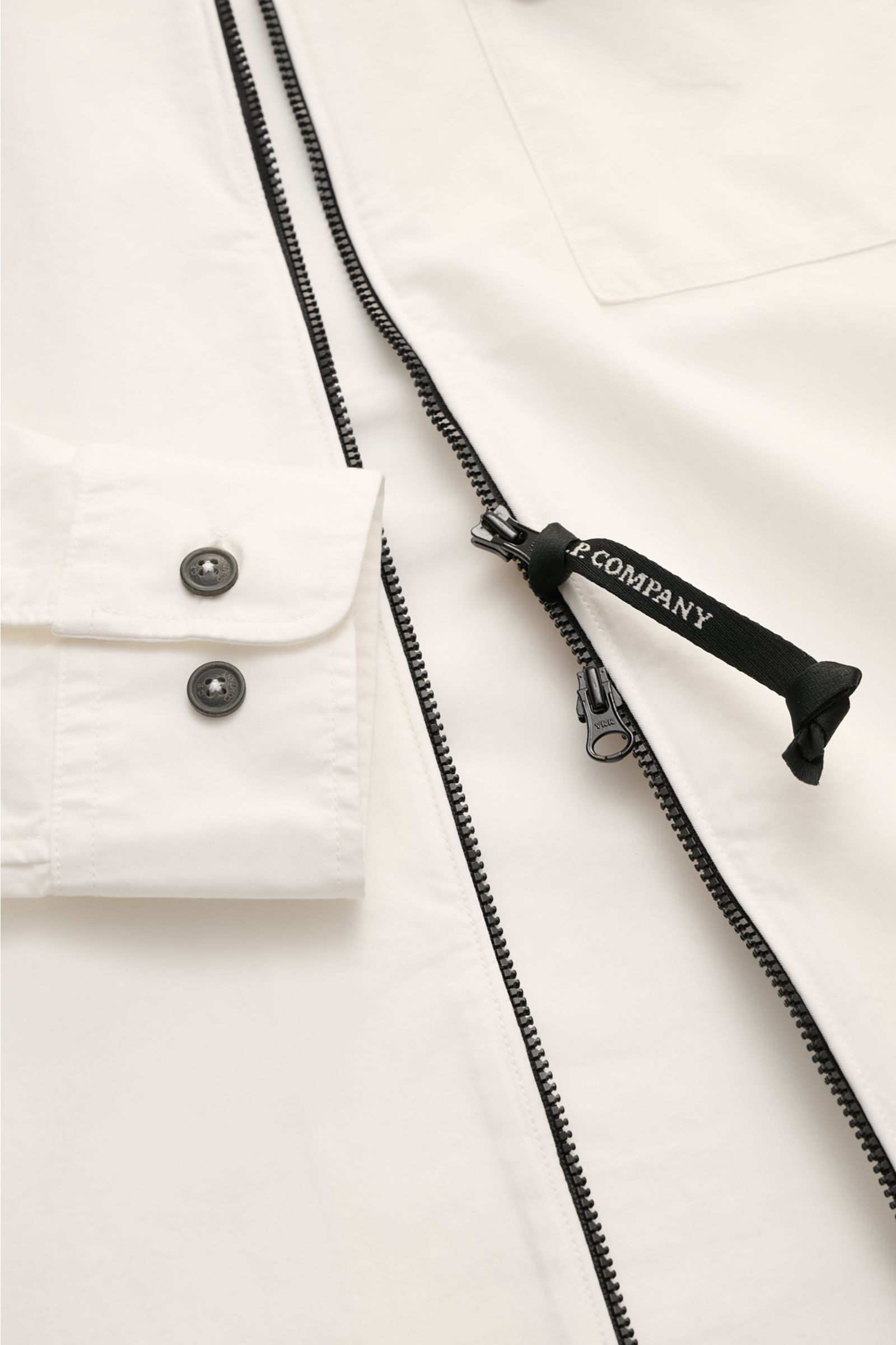 Close-up view of the C.P. Company Overshirt offwhite showing slim fit, two-way black zipper with logo pull, cuff with two buttons, and chest pocket flap.