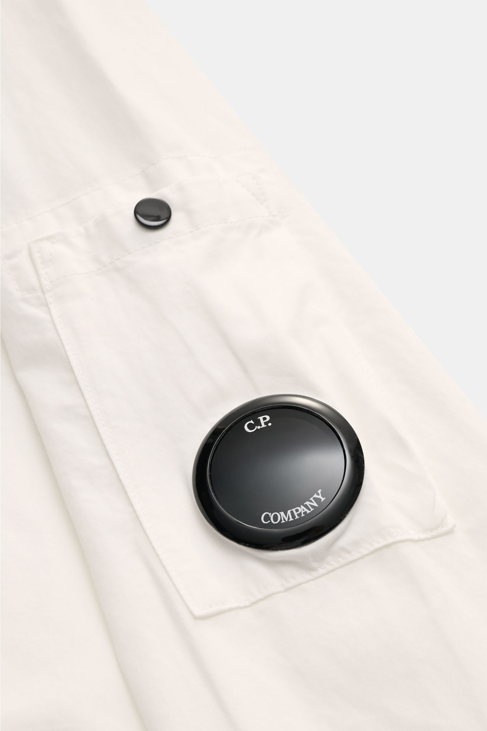 Close-up front detail of C.P. Company Overshirt offwhite showing pocket with black logo lens, photographed from a top angle. Lässiges Layering-Piece: Das Overshirt von C.P. COMPANY ist der ideale Begleiter für urbane Frühlingslooks. reine Baumwolle, leich