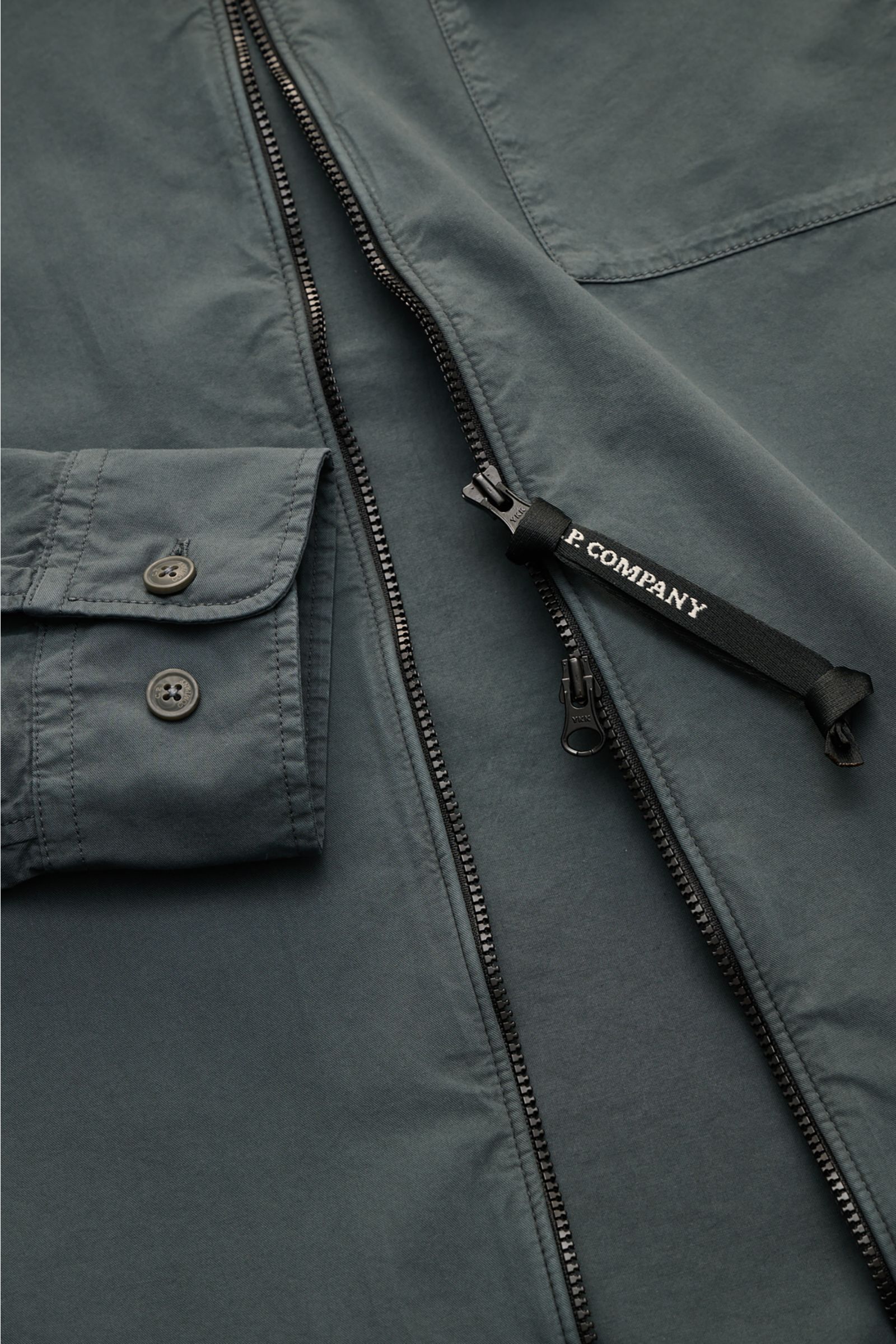 Close-up front view of the C.P. Company Overshirt dunkelgrau showing slim fit, pure cotton, lightweight fabric, two-way zipper with logo pull, cuffed sleeves, chest pocket with flap, and sleeve logo detail. Lässiges Layering-Piece: Das Overshirt von C.P. 
