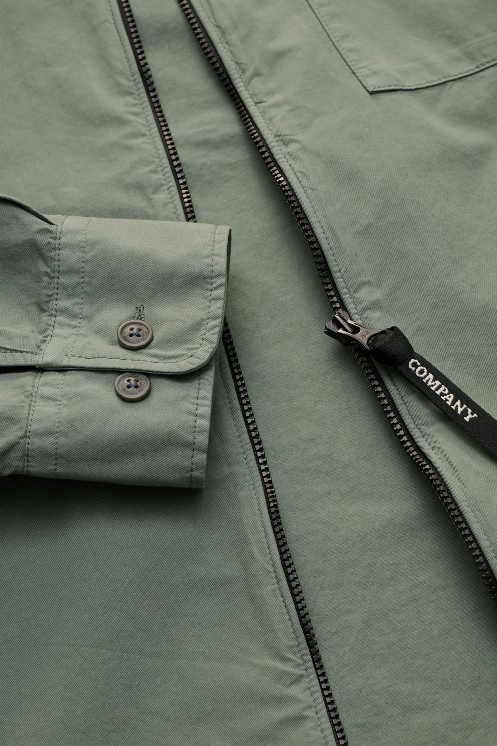 Close-up front view of the C.P. Company Overshirt salbei in slim fit, lightweight pure cotton with two-way zipper, chest pocket with flap, sleeve logo detail, cuff buttons, and turn-down collar, perfect as a casual layering piece for urban spring looks.