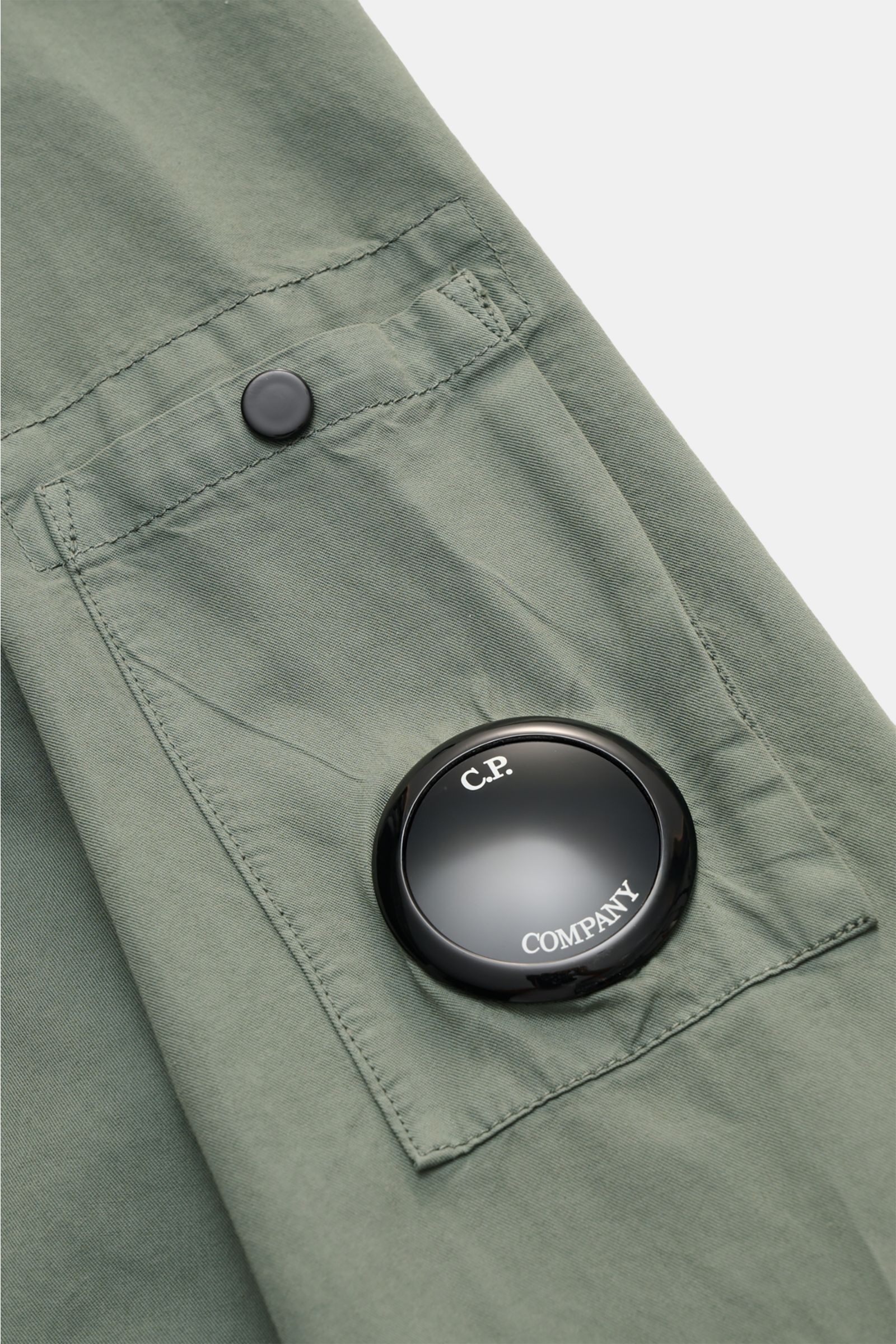 Close-up view of the C.P. Company Overshirt salbei sleeve showing a buttoned chest pocket and a black logo detail badge on pure cotton slim fit fabric. Lässiges Layering-Piece: Das Overshirt von C.P. COMPANY ist der ideale Begleiter für urbane Frühlingslo