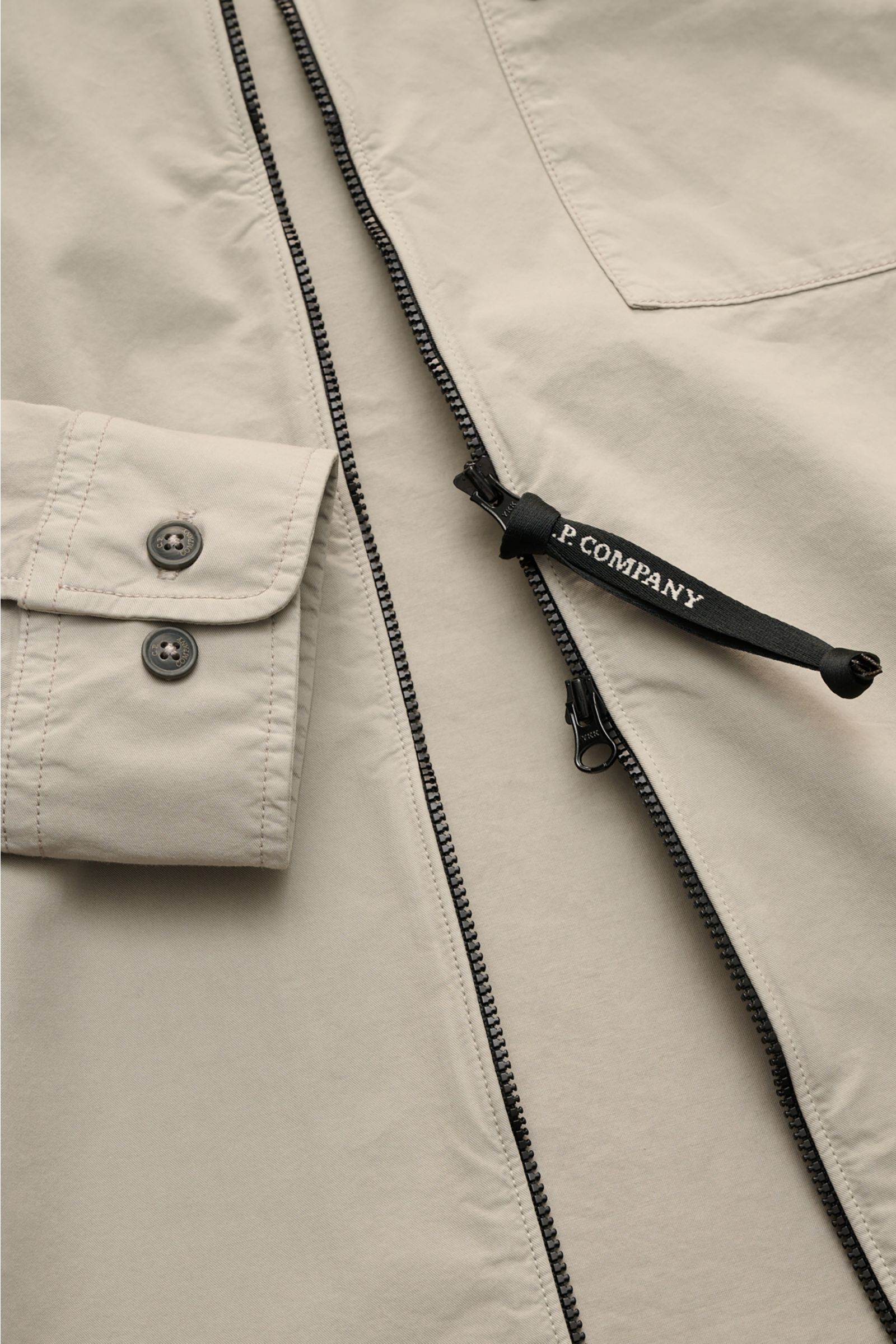 Close-up top view of the C.P. Company Overshirt sand showing slim fit, two-way zipper with logo pull, cuff with buttons, and chest pocket with flap.