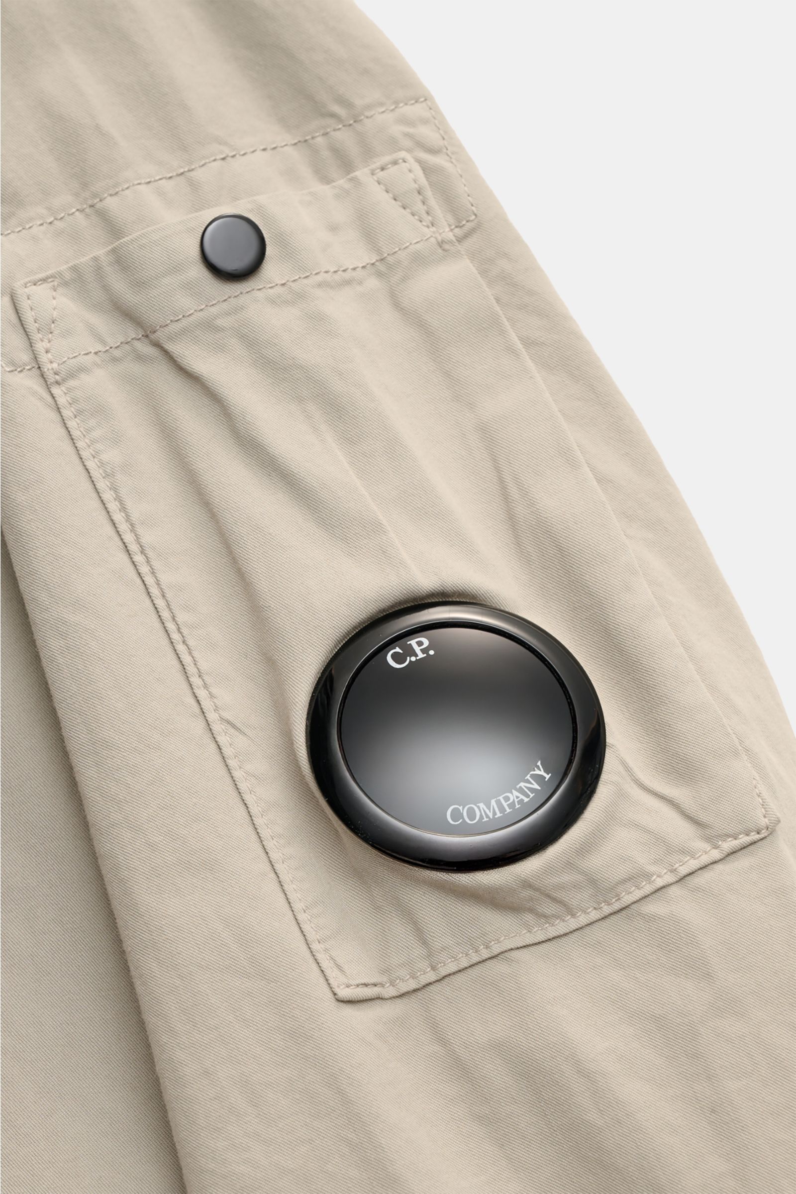 Close-up from above of the sand-colored C.P. Company Overshirt sleeve pocket with button flap and black logo lens detail, made of pure cotton. Lässiges Layering-Piece: Das Overshirt von C.P. COMPANY ist der ideale Begleiter für urbane Frühlingslooks. Rein