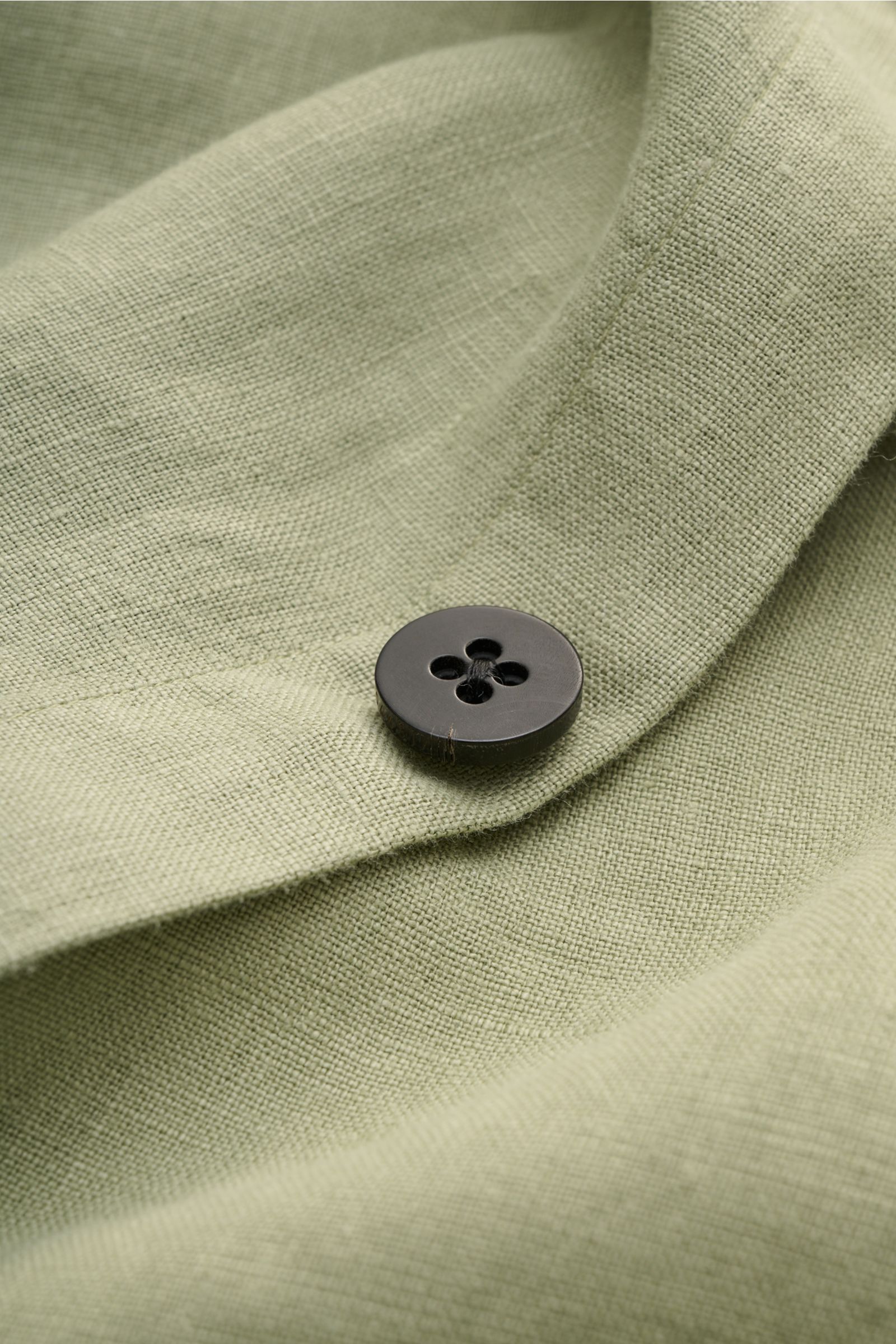Nahaufnahme aus schräger Perspektive des salbeigrünen Leinens mit Knopf, Detail des 04651/ A trip in a bag Overshirt 'Linen Jacket' salbei, reines Leinen, weicher Griff, Slim Fit, Fünf-Knopf-Leiste, Umlegekragen, aufgesetzte Fronttaschen und Innentasche.