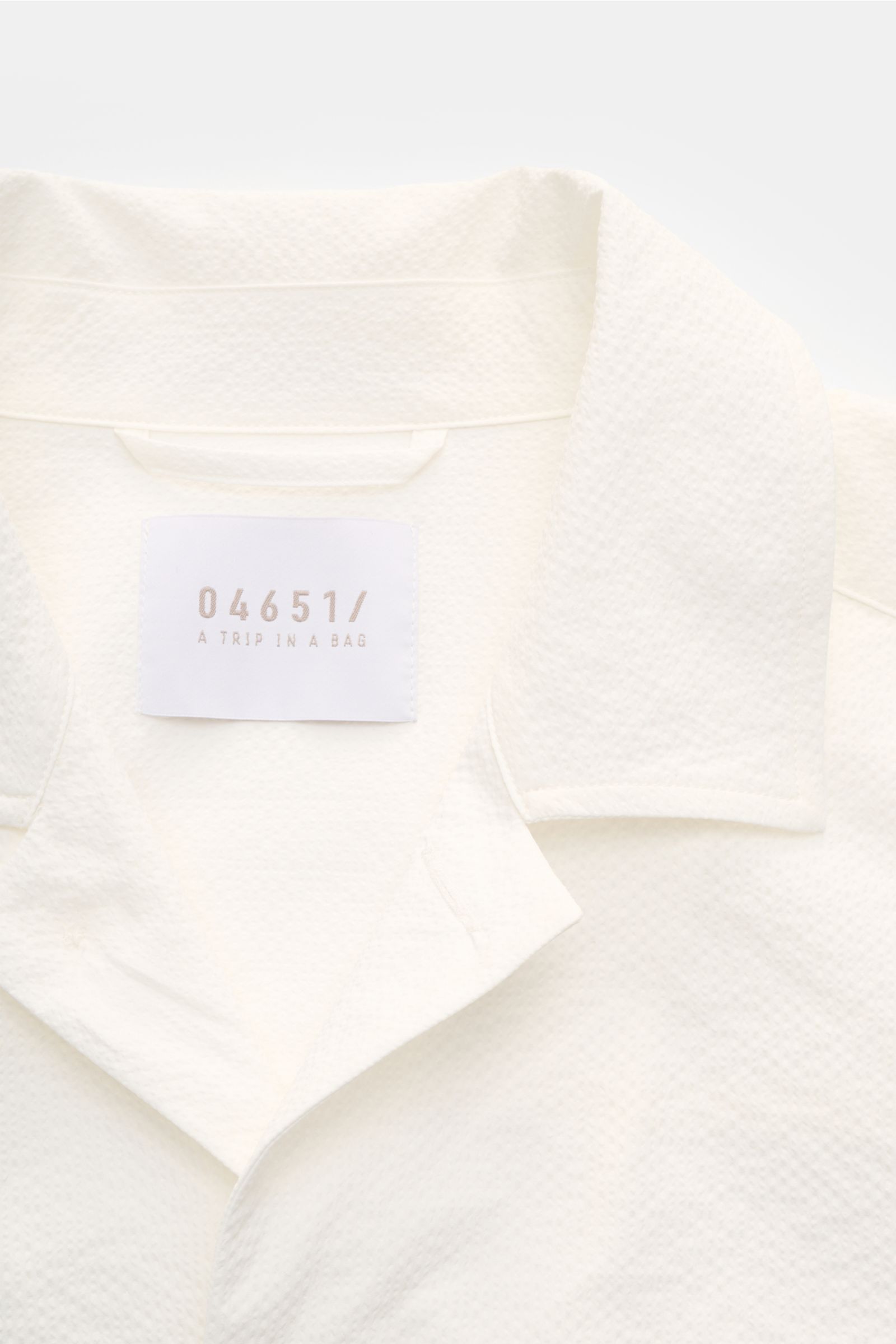 Close-up front top view of the 04651/ A trip in a bag Overshirt 'Technical Seersucker' off-white showing textured microfibre fabric, turn-down collar, and label.

Textured holiday essential: This seersucker overshirt by 04651/ A TRIP IN A BAG is a vers