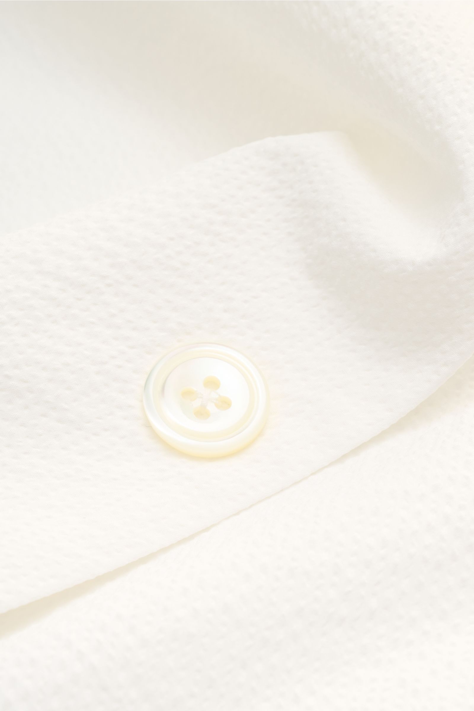 Close-up front view of the off-white 04651/ A trip in a bag Overshirt 'Technical Seersucker' showing a textured seersucker fabric and a four-hole button. Textured holiday essential: This seersucker overshirt by 04651/ A TRIP IN A BAG is a versatile piece 