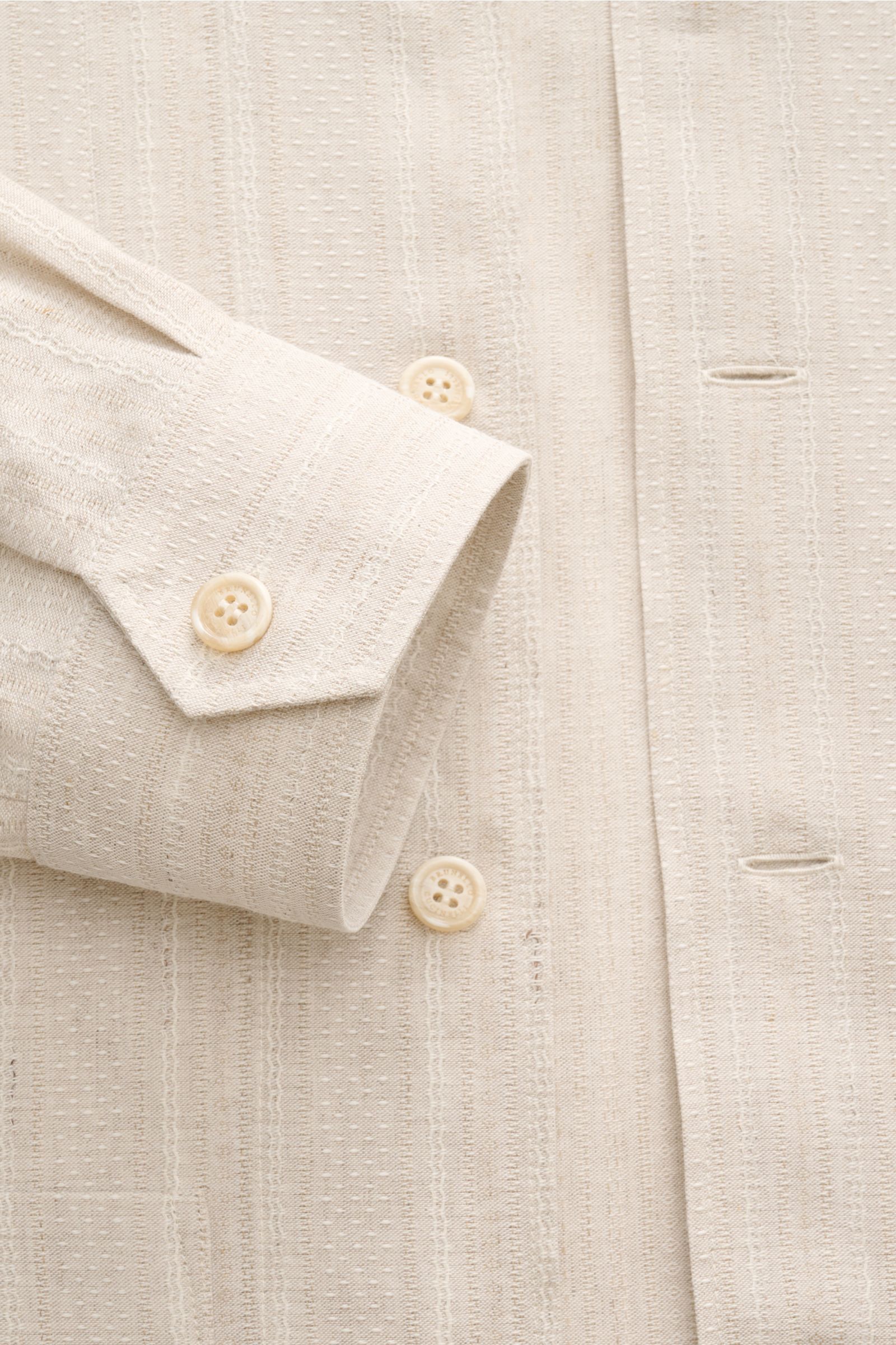 Close-up front view of the beige Brunello Cucinelli Overshirt showing slim fit, linen-cotton mix, seven-button placket, cuff with buttons, unconstructed design, and textured fabric.

Zeitloses Layering-Piece: Das Overshirt von BRUNELLO CUCINELLI ergänz