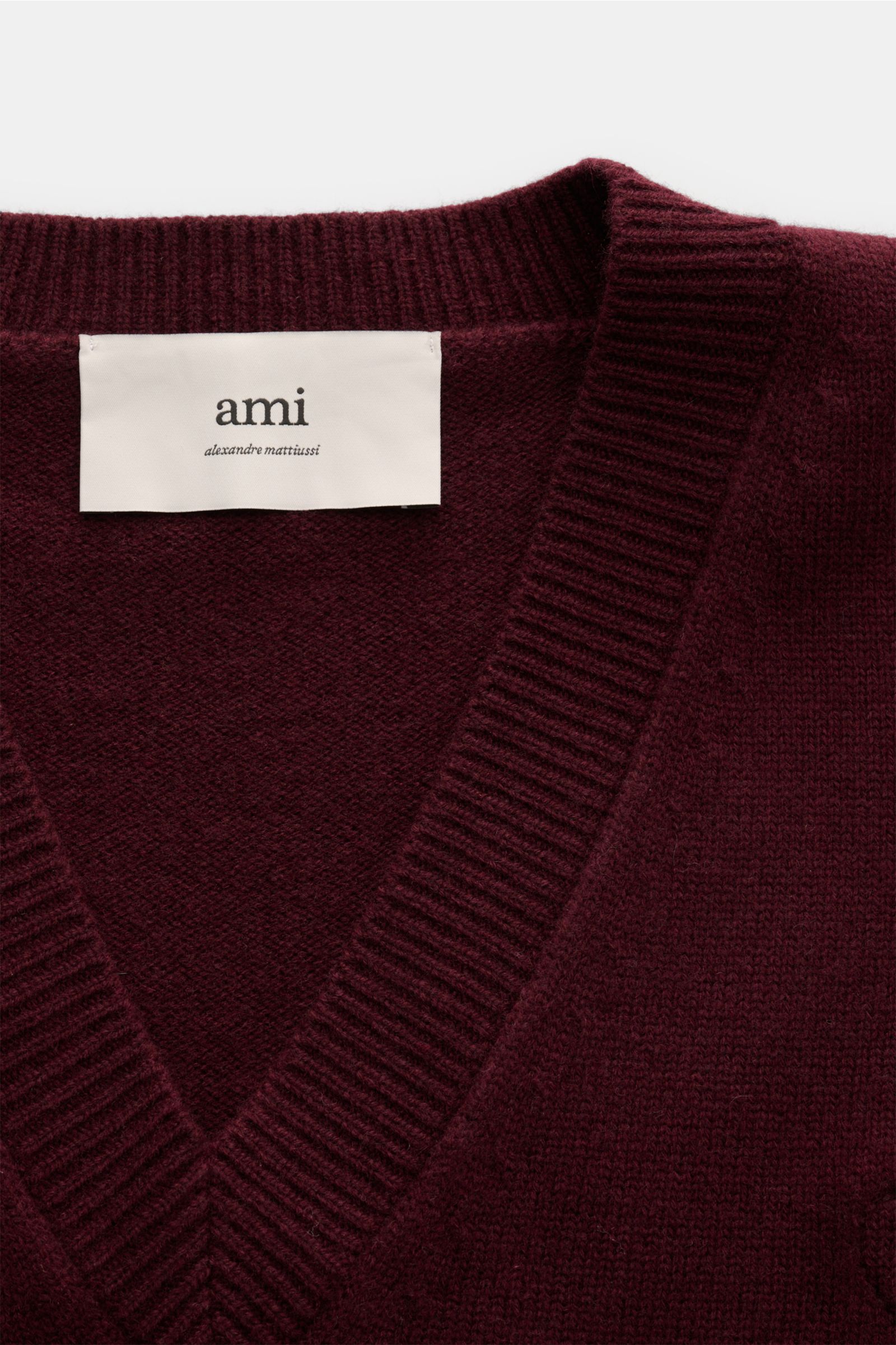 Nahaufnahme des Ami Paris V-Neck Pullovers bordeaux aus Wolle und Cashmere mit weichem Griff, mittelfeinem Strick, breiten Rippbündchen und abgestepptem 'Ami'-Logo, fotografiert von oben auf den V-Neck.
