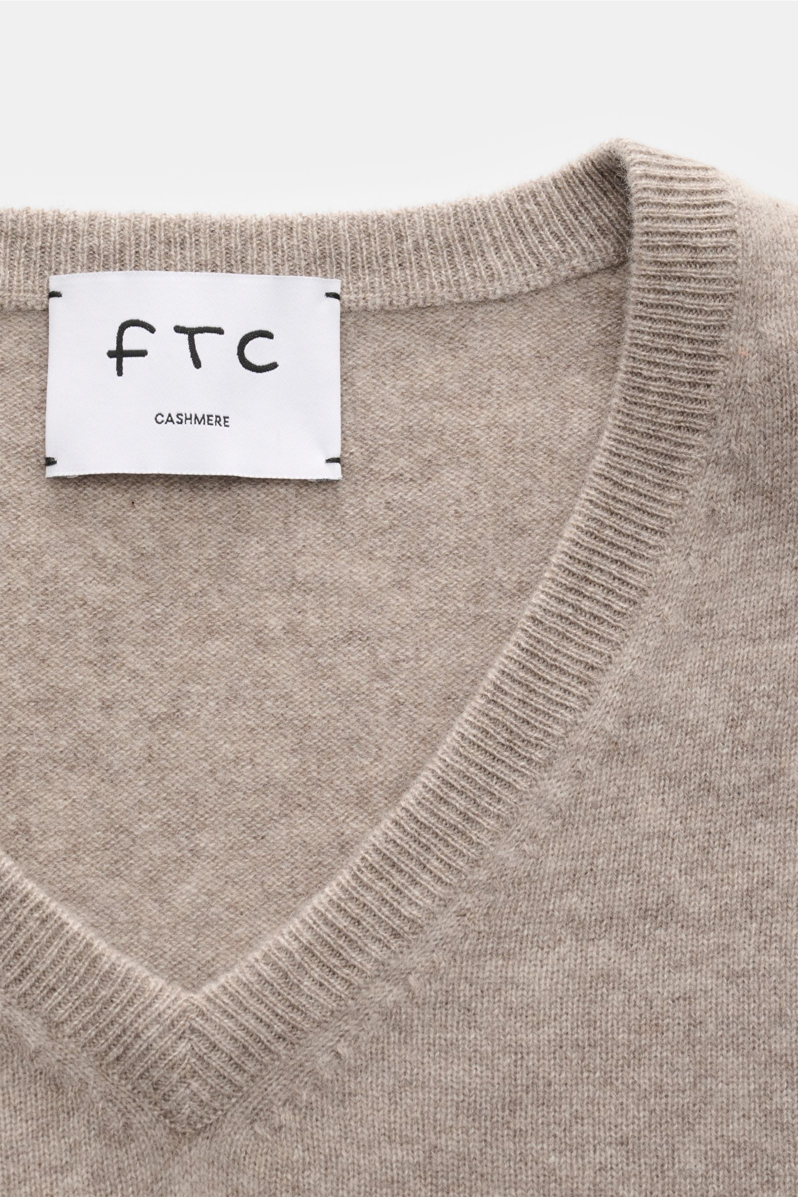 Close-up front view of the FTC Cashmere V-Neck Pullover taupe showing the fine knit texture, V-neckline, and ribbed collar with brand label.

Der Pullover mit V-Ausschnitt ist ein Klassiker der Herrenmode, der in keinem Kleiderschrank fehlen darf – aus