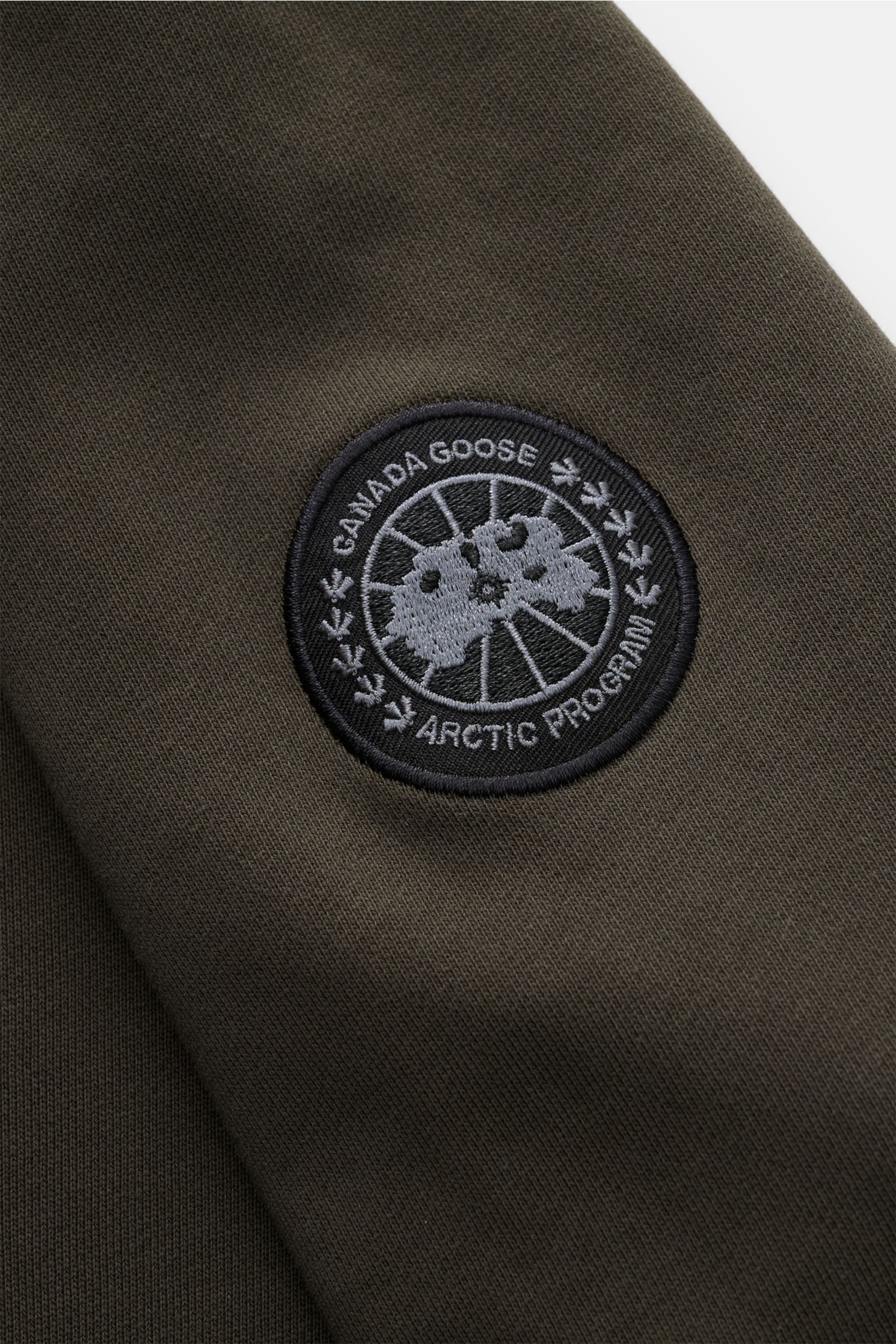 Alt tag (125 characters max): Close-up of Canada Goose Kapuzenpullover 'Huron' beige sleeve showing black and gray embroidered CANADA GOOSE patch.

Description: Urbaner Style trifft Komfort – der Kapuzenpullover 'Huron' von CANADA GOOSE verbindet angen